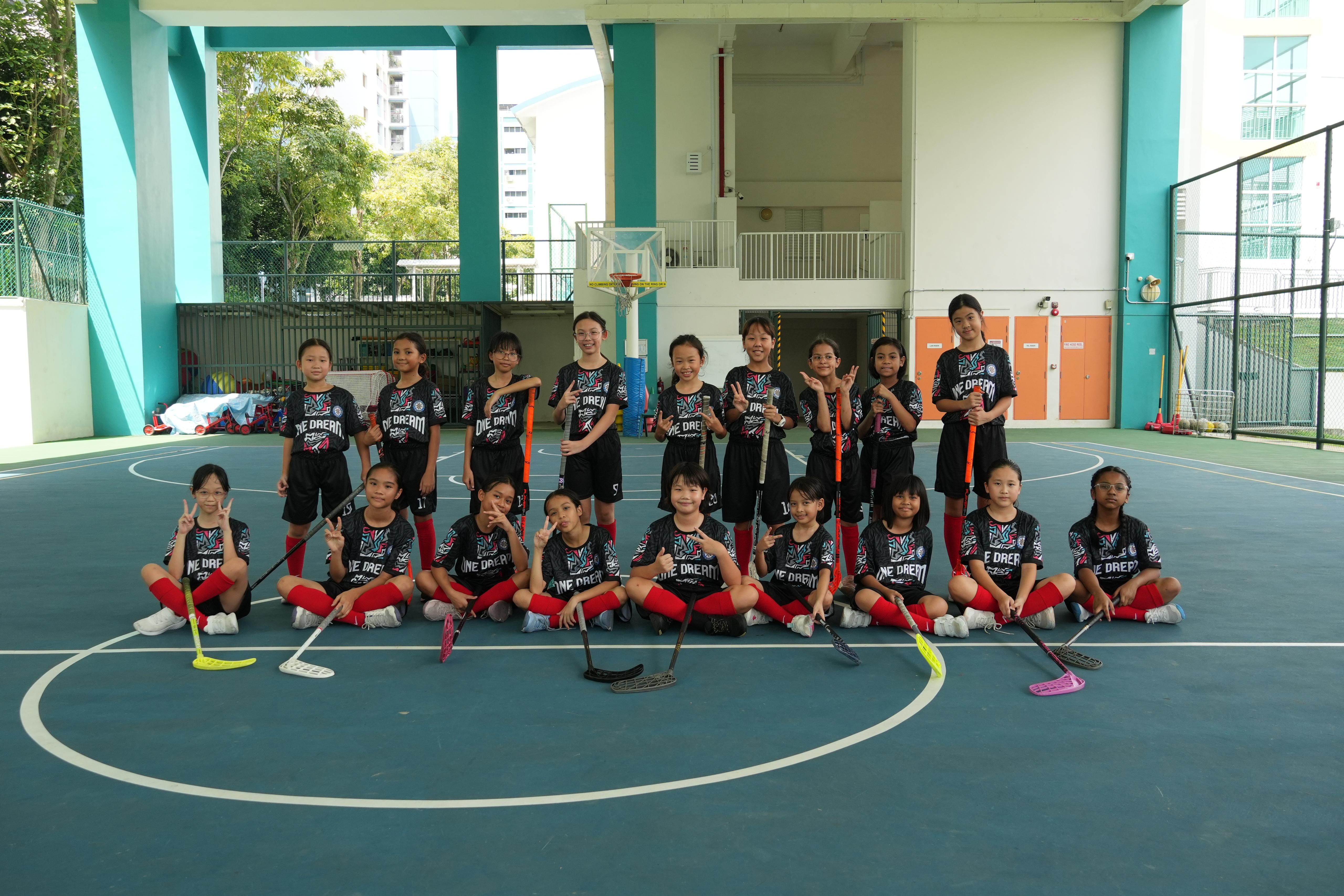 A group of young girls in black uniforms pose with floorball sticks on an outdoor court.