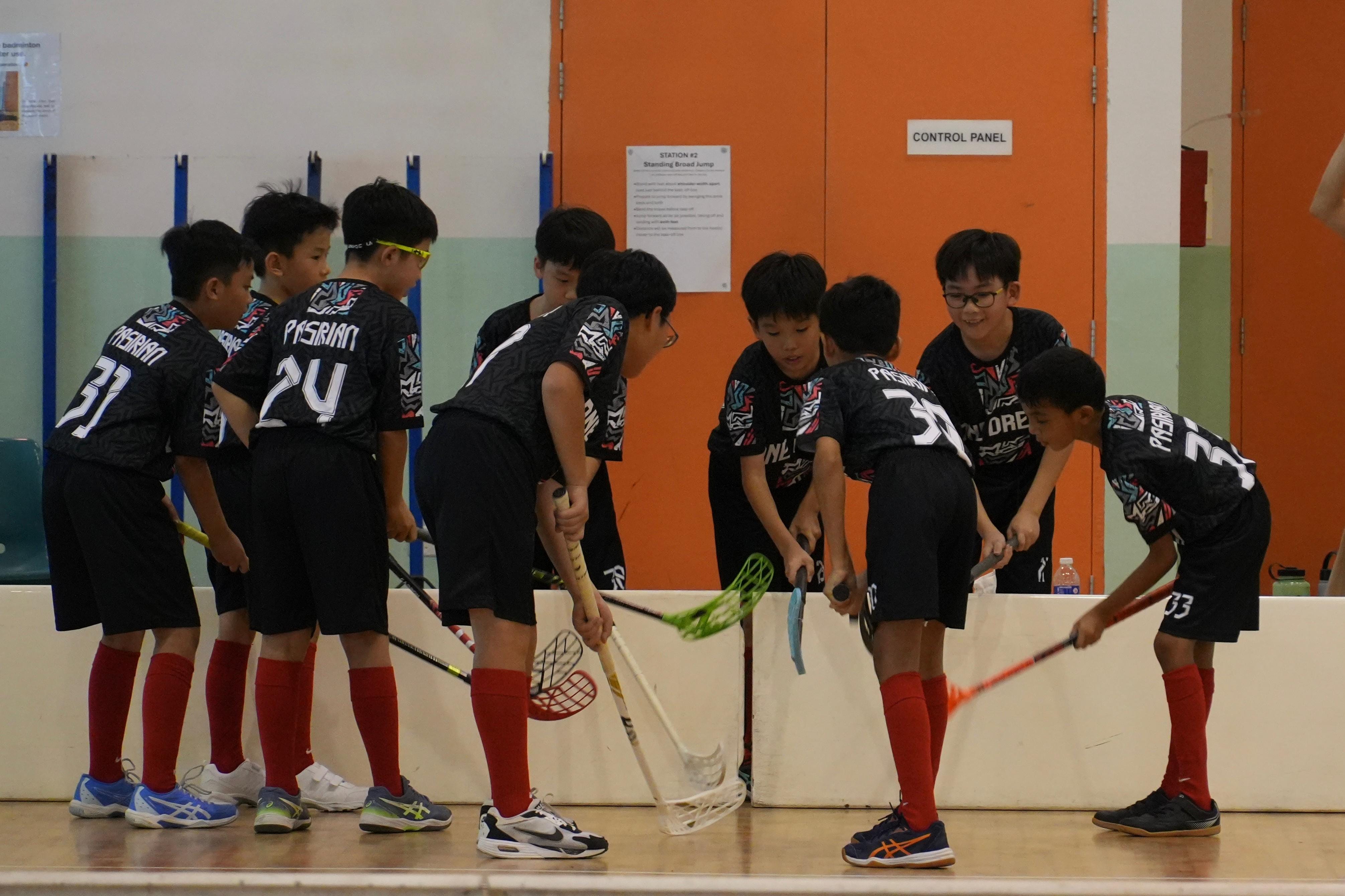 A group of young boys in black jerseys and red socks play indoor floorball, holding sticks in a gymnasium.