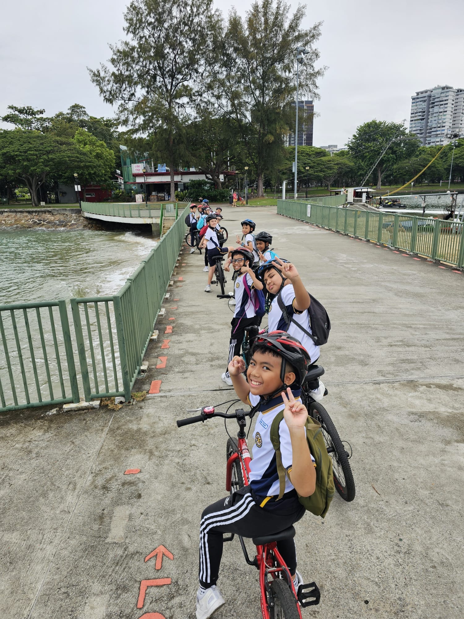 A group of children wearing helmets and backpacks pose on bicycles along a waterfront path lined with green railings.