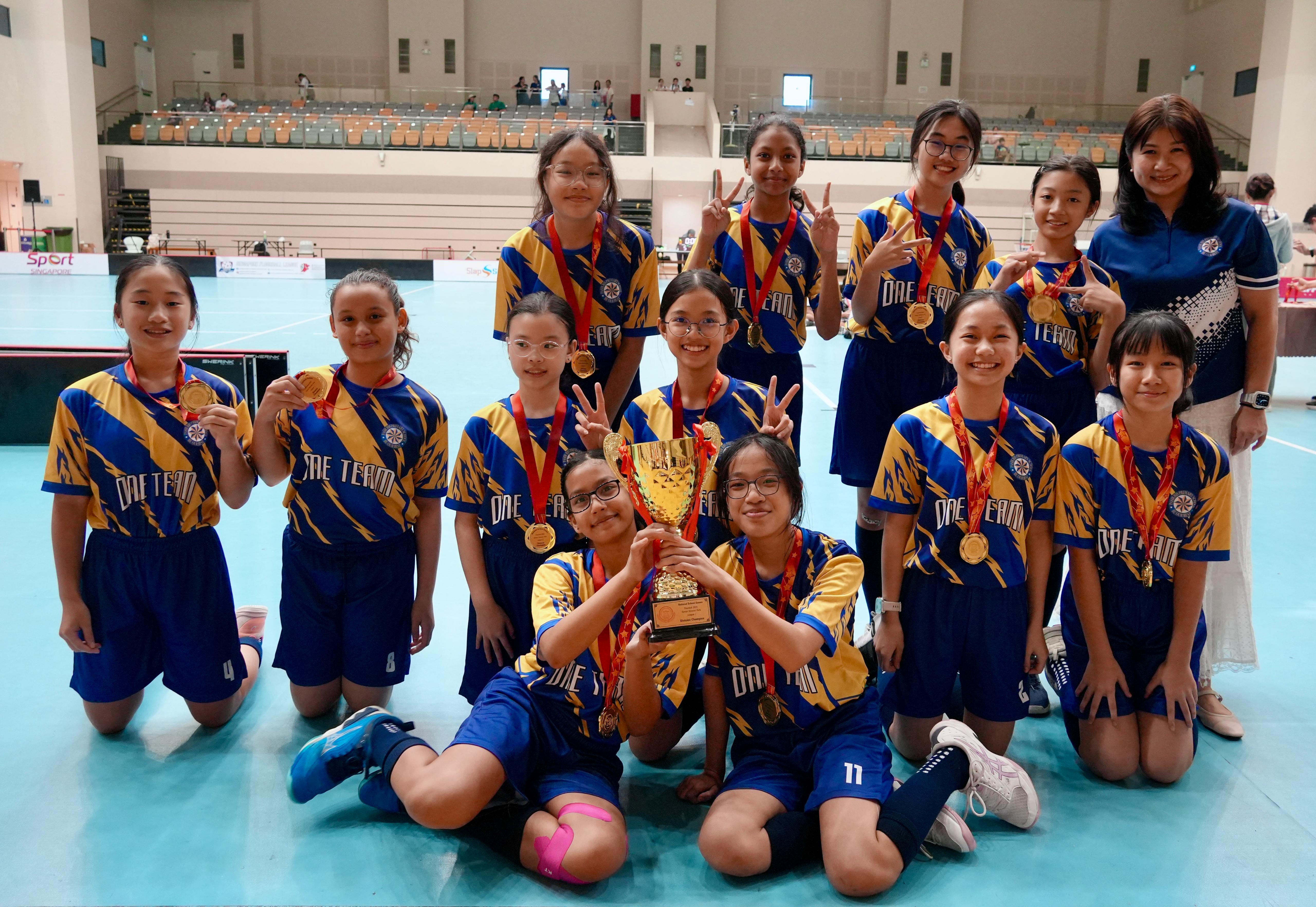 A girls sports team in blue and gold uniforms poses with gold medals and a trophy inside an indoor arena.