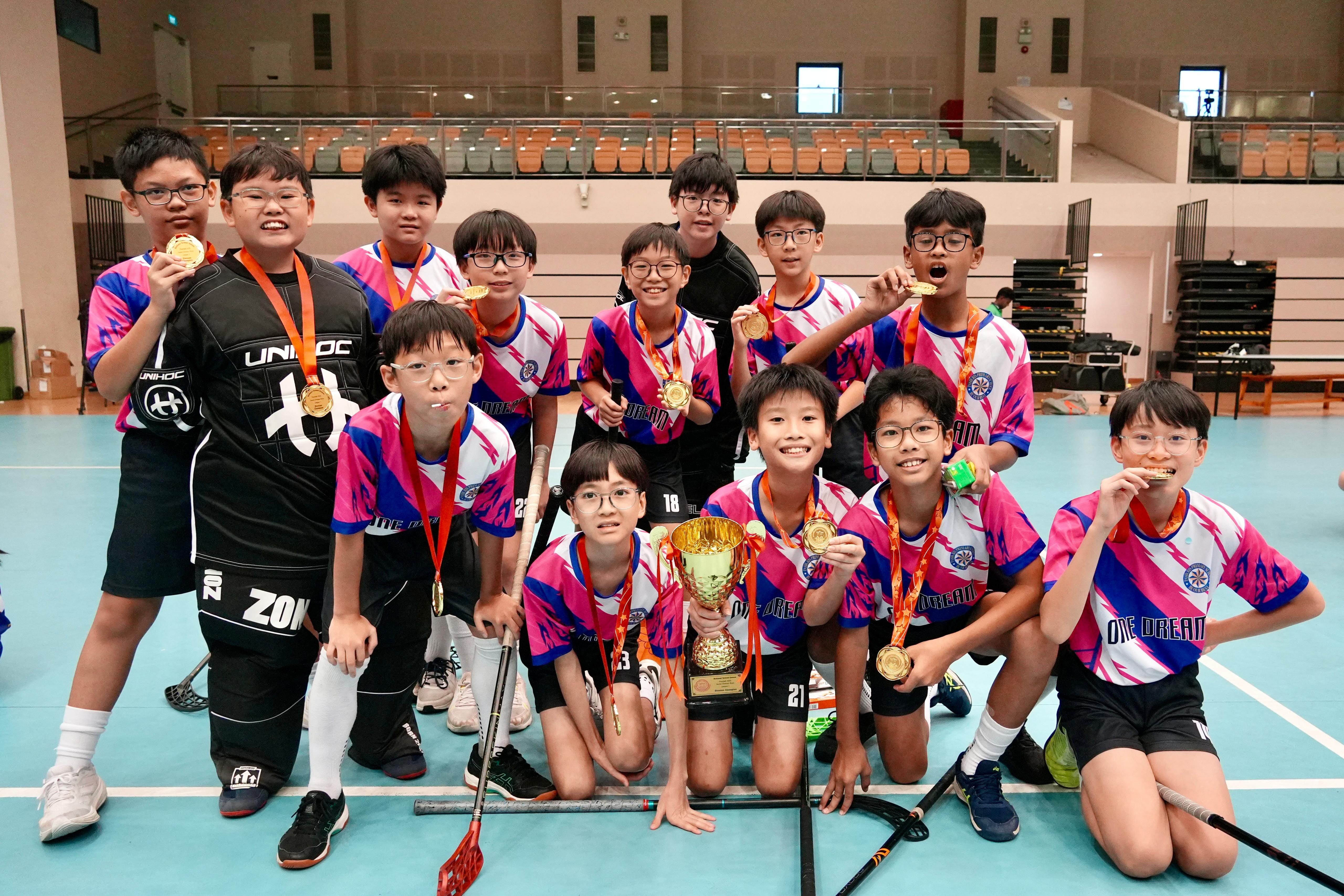 A youth floorball team in pink and blue jerseys poses with gold medals and a trophy inside an indoor sports hall.