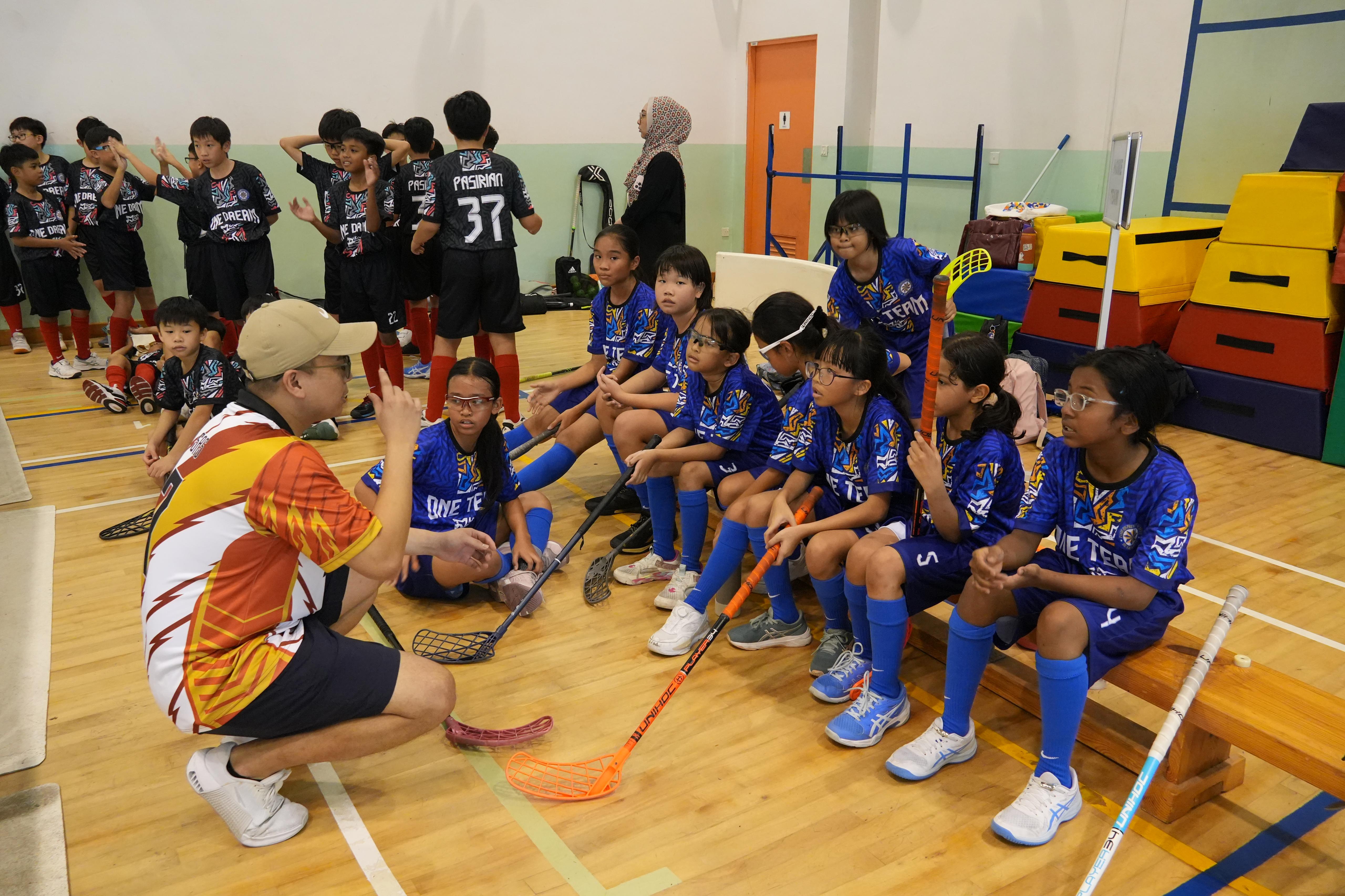 A coach in a cap talks to young girls in blue uniforms holding floorball sticks inside a gymnasium.