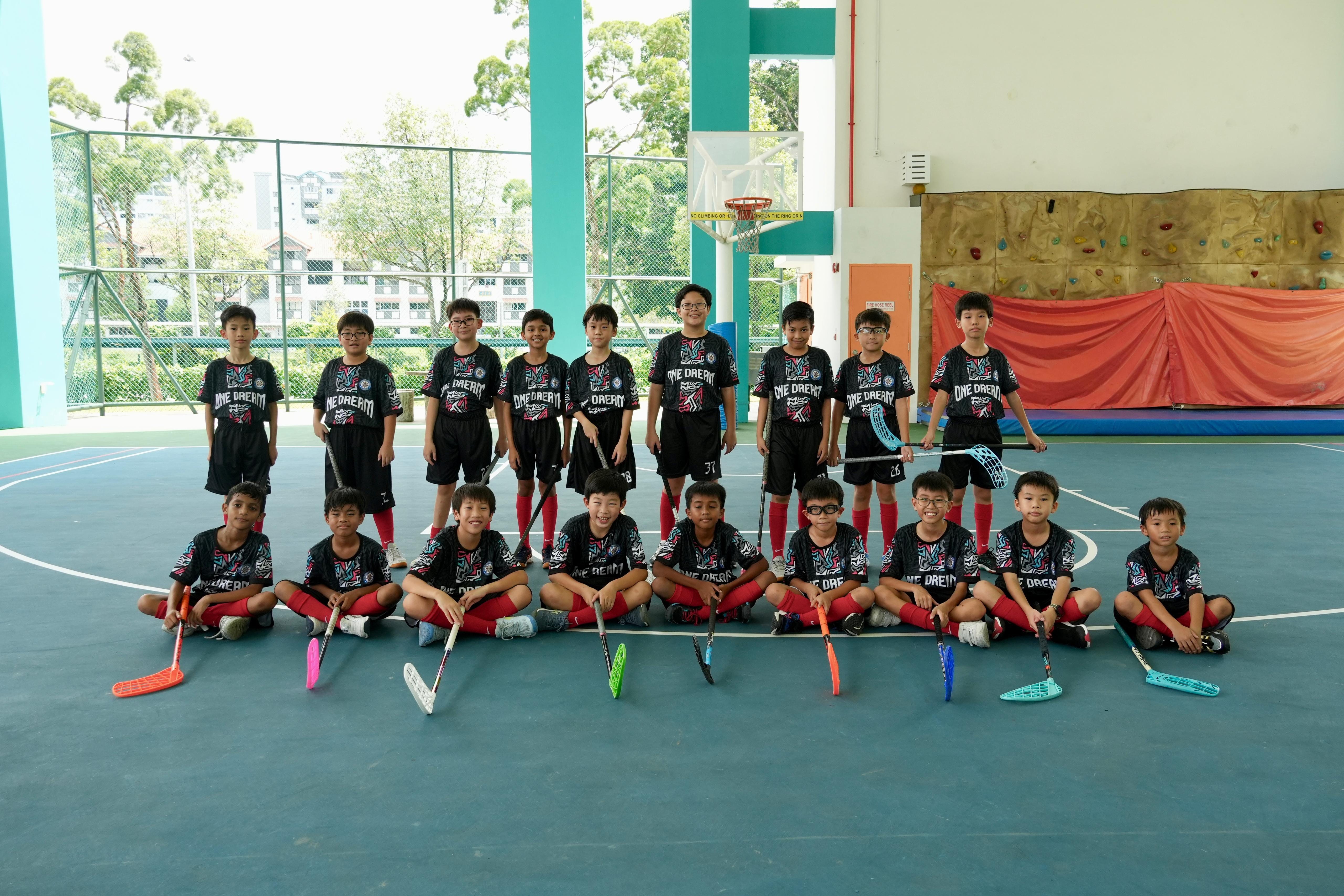A youth floorball team in black uniforms poses with colorful sticks on an indoor court.