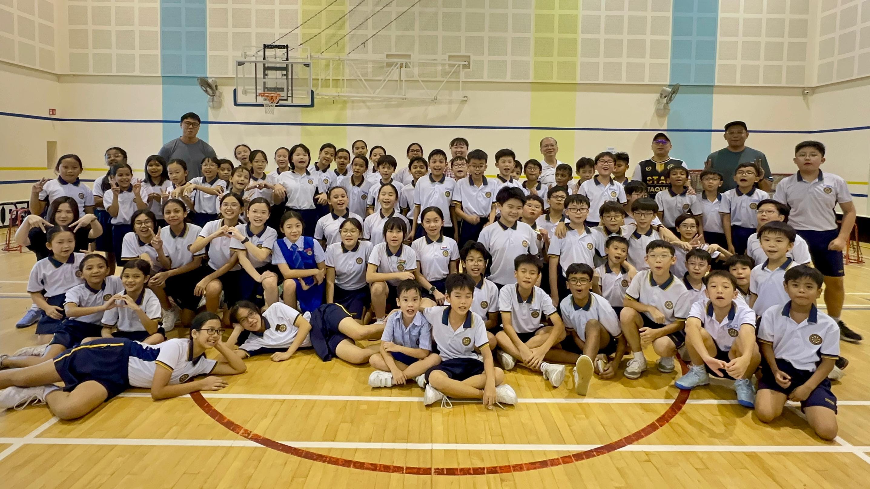 Large group of children in white and navy school uniforms posing with adults in an indoor basketball gymnasium.