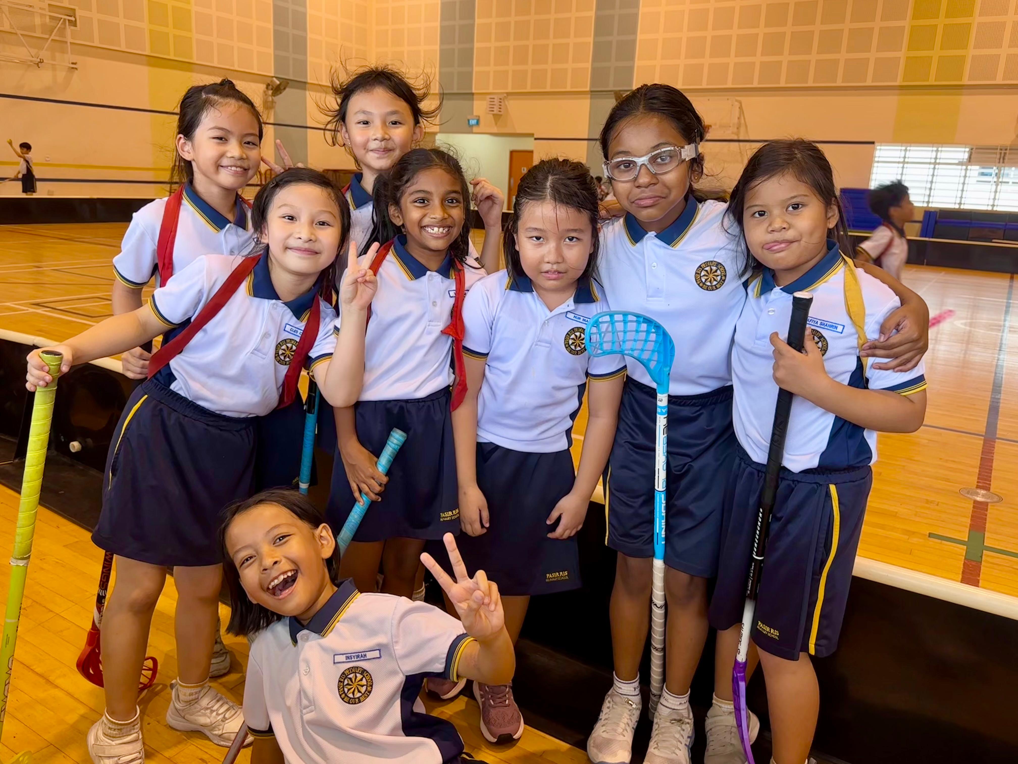 Eight young students in matching uniforms smile and pose together in a gymnasium, holding floorball sticks.