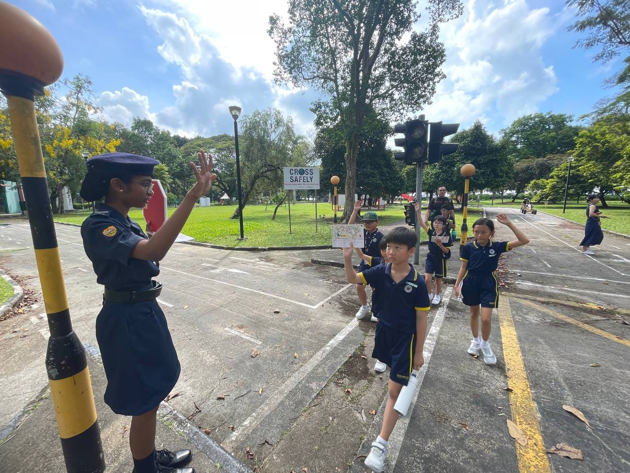 A uniformed officer teaches young students in navy uniforms to raise their hands while crossing a road safely outdoors.