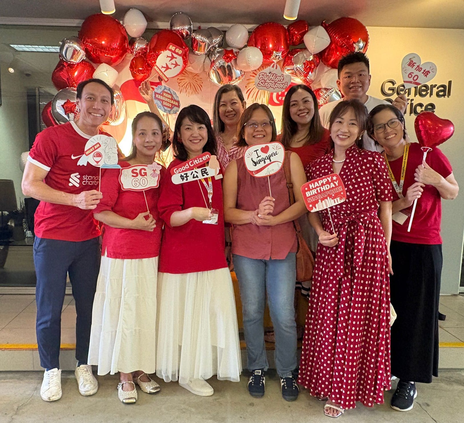 Nine people in red outfits pose with Singapore SG60 National Day signs before red and white balloons.