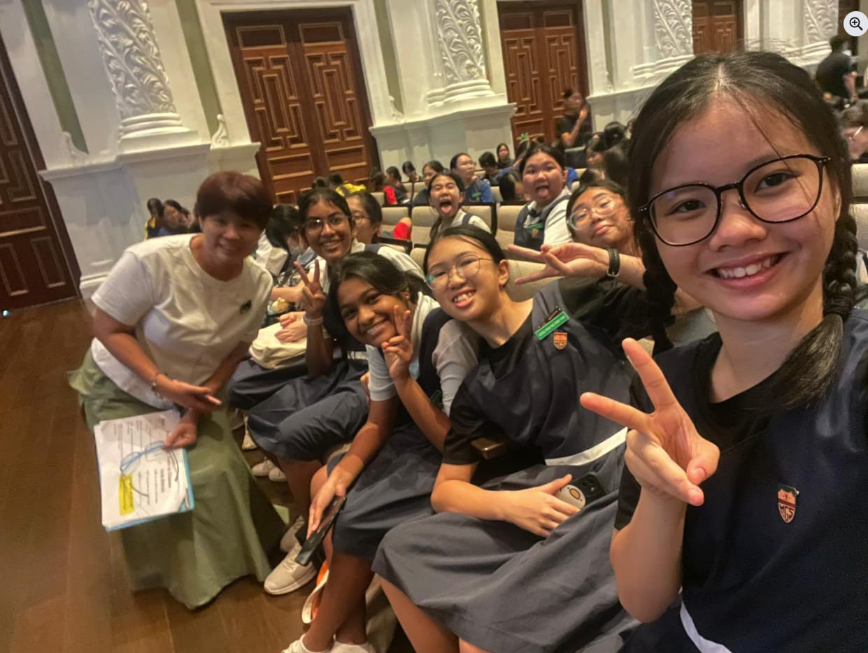 A group of smiling schoolgirls in navy uniforms pose with peace signs in an ornate hall with wooden doors.