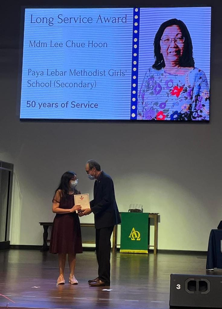 Two masked people exchange a certificate on stage below a screen showing a Long Service Award for 50 years.