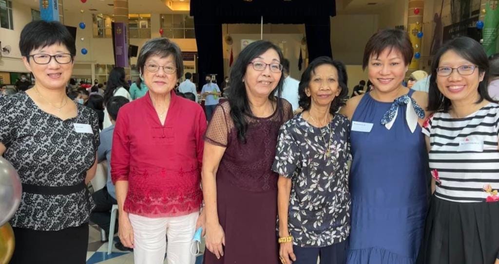 Six women smiling and posing together at an indoor event with balloons and a crowd in the background.