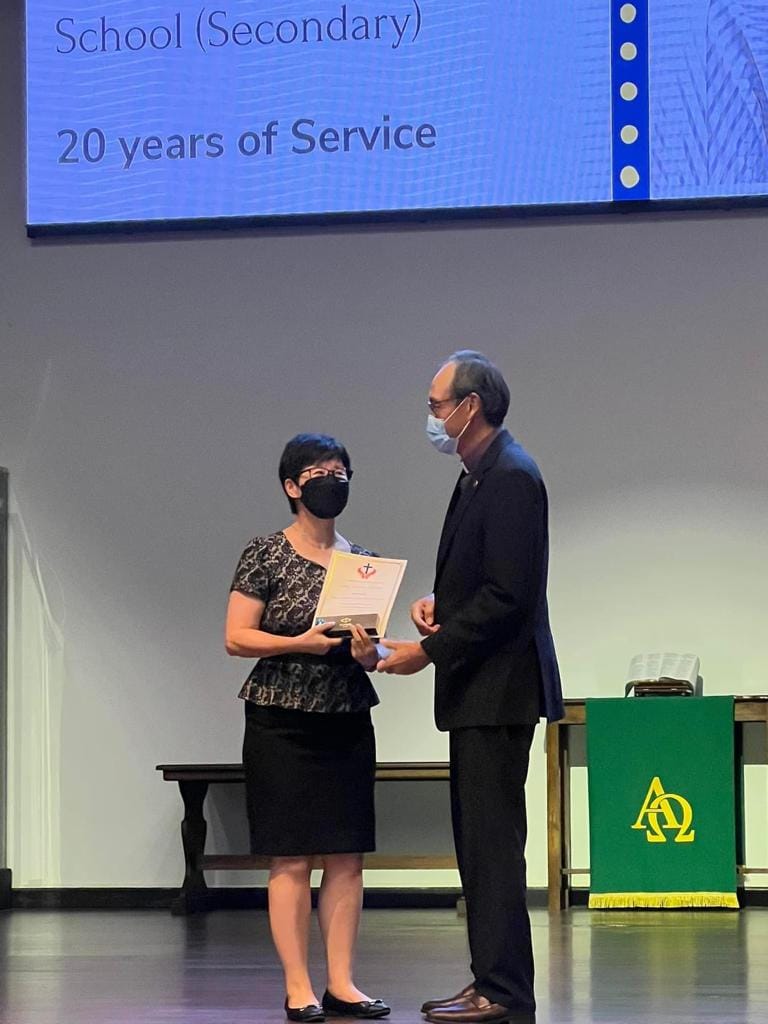 Two masked people on stage exchange a certificate at a 20 years of service award ceremony.