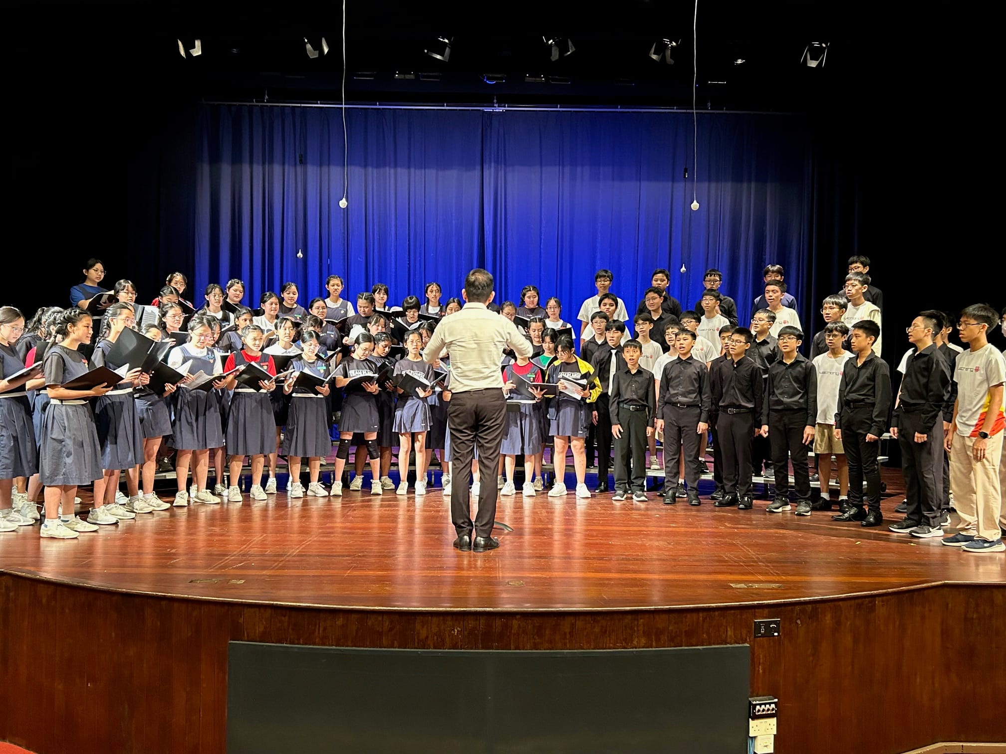 A conductor stands with back to camera leading a large student choir on a wooden stage with blue curtains.