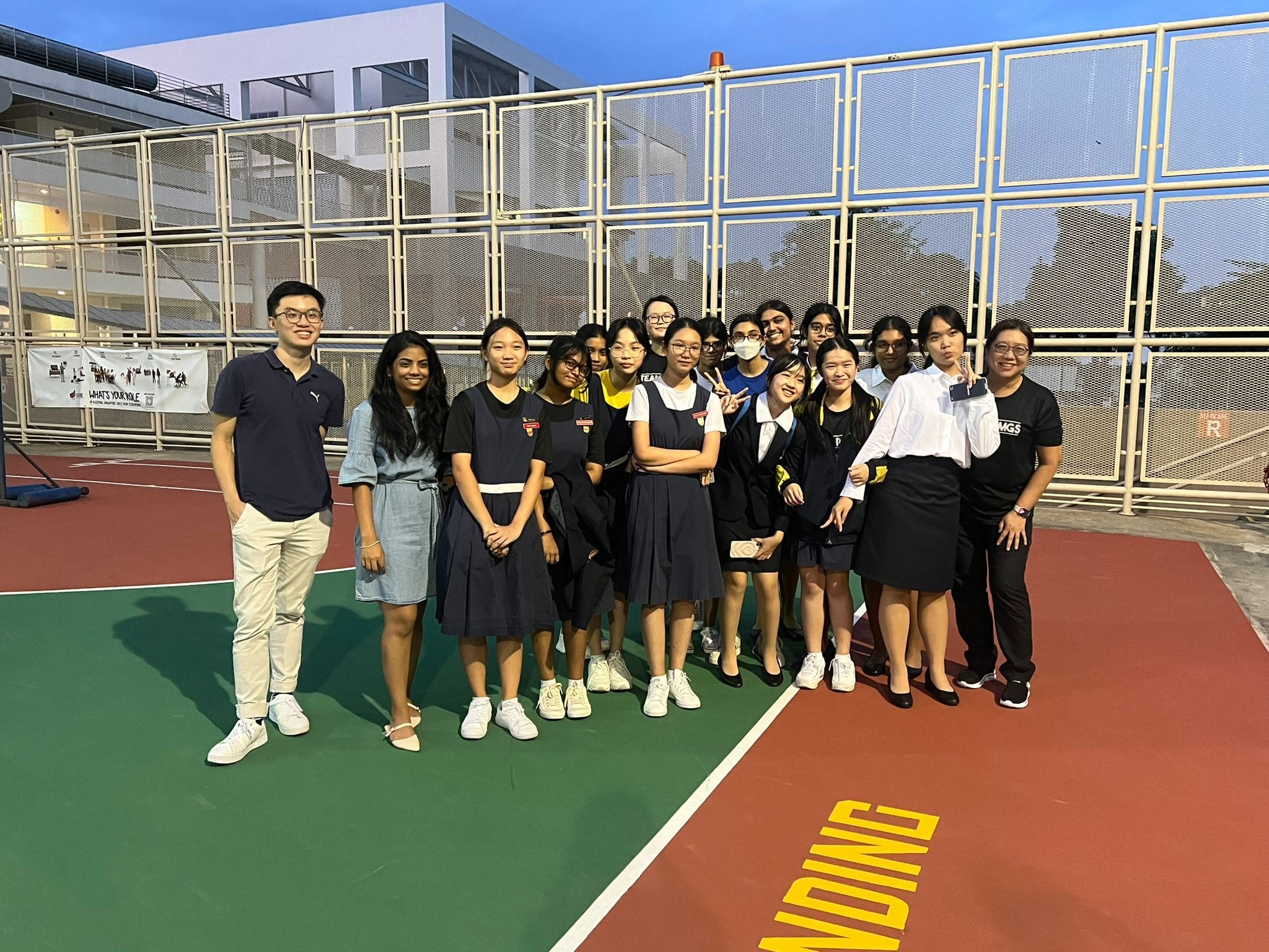 A group of about 15 students and two adults posing together on an outdoor sports court near a fenced building.