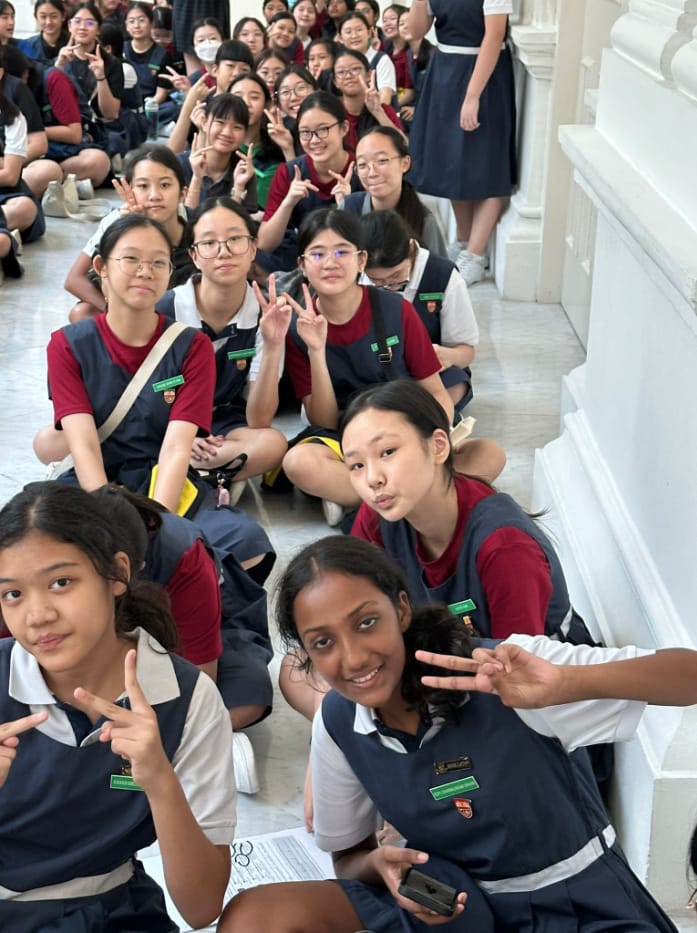 A large group of schoolgirls in navy and red uniforms sit on a floor, smiling and making peace signs.