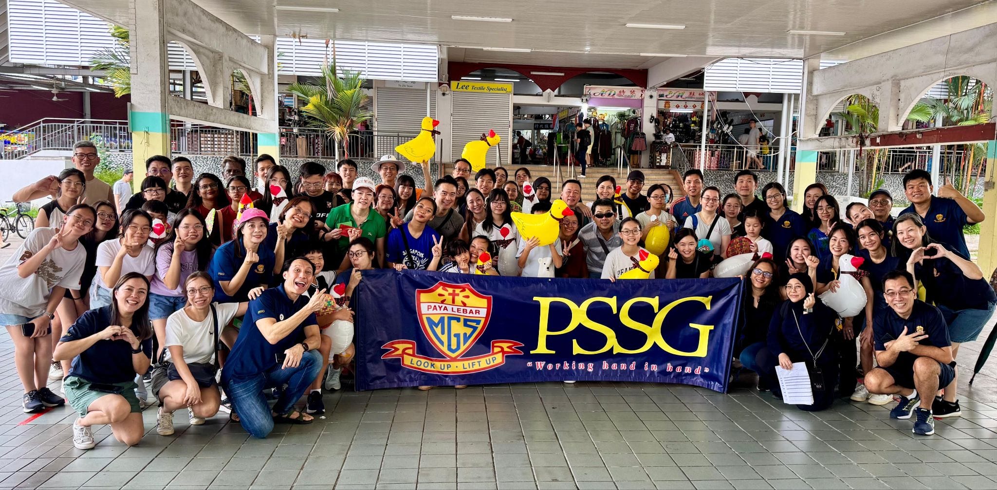 Large group posing outdoors holding a navy banner reading Paya Lebar MGS PSSG Working hand in hand.