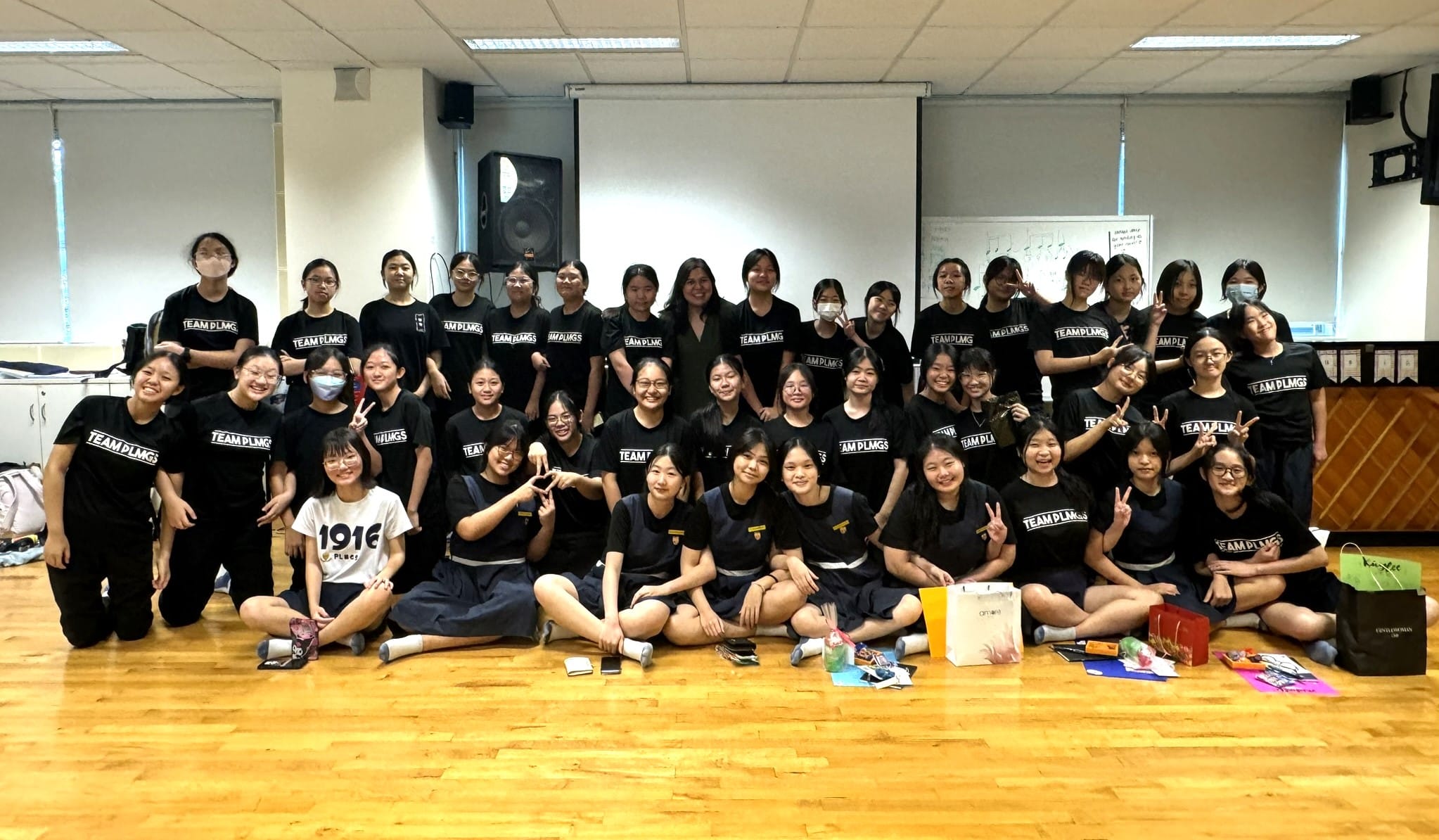 Large group of young women posing indoors, most wearing black Team PLMGS shirts, smiling and making peace signs.