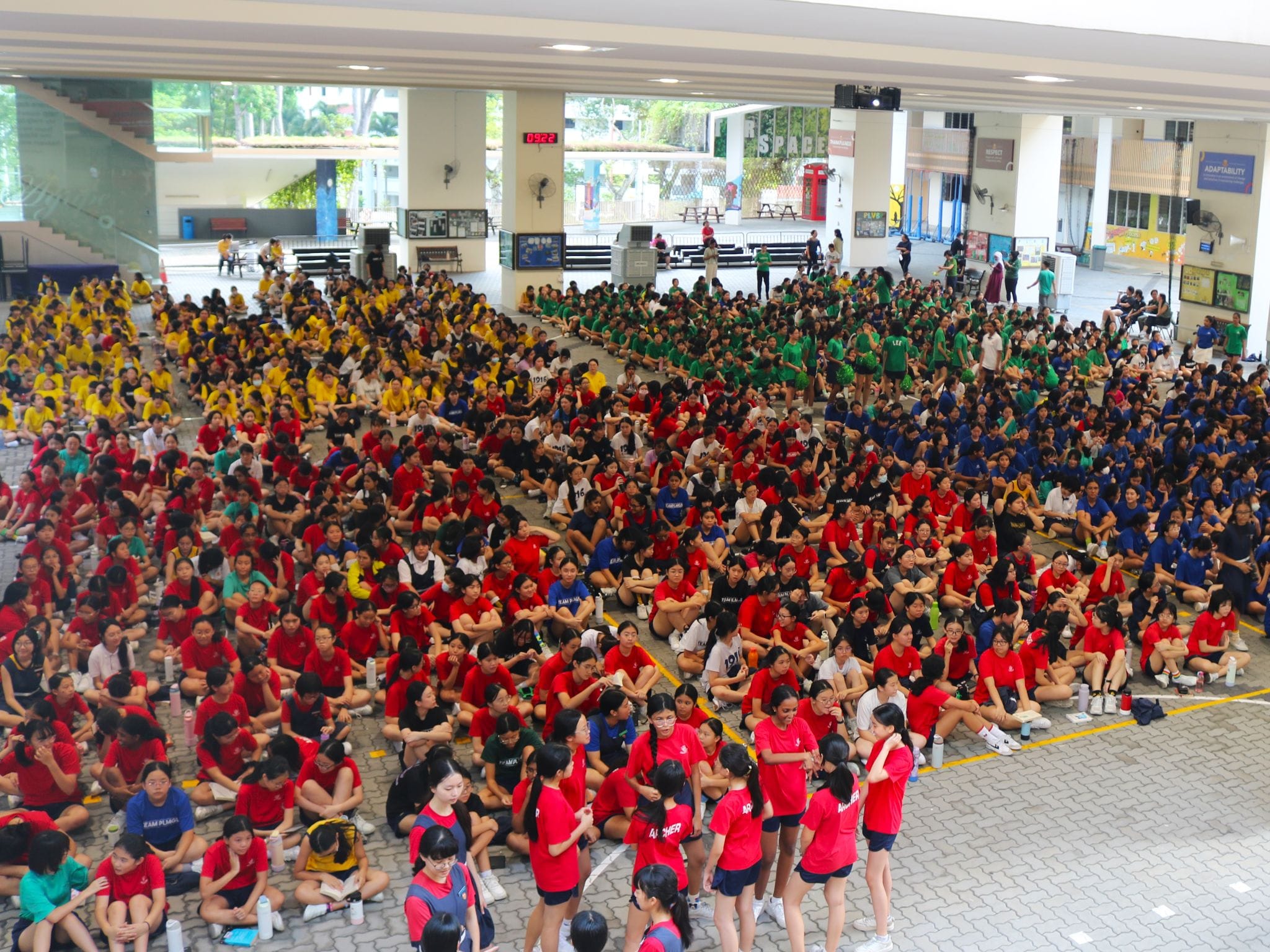 Large crowd of students in red, yellow, green, and blue shirts sitting on the ground in a covered school courtyard.