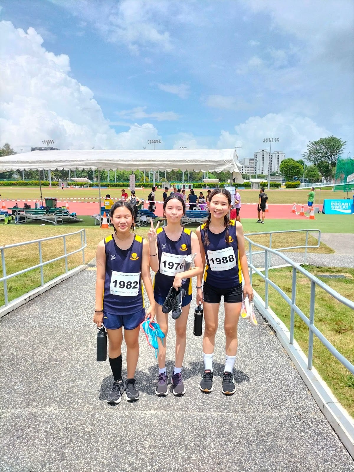Three young female athletes in navy uniforms with race bibs 1982, 1976, and 1988 pose at an outdoor track meet.