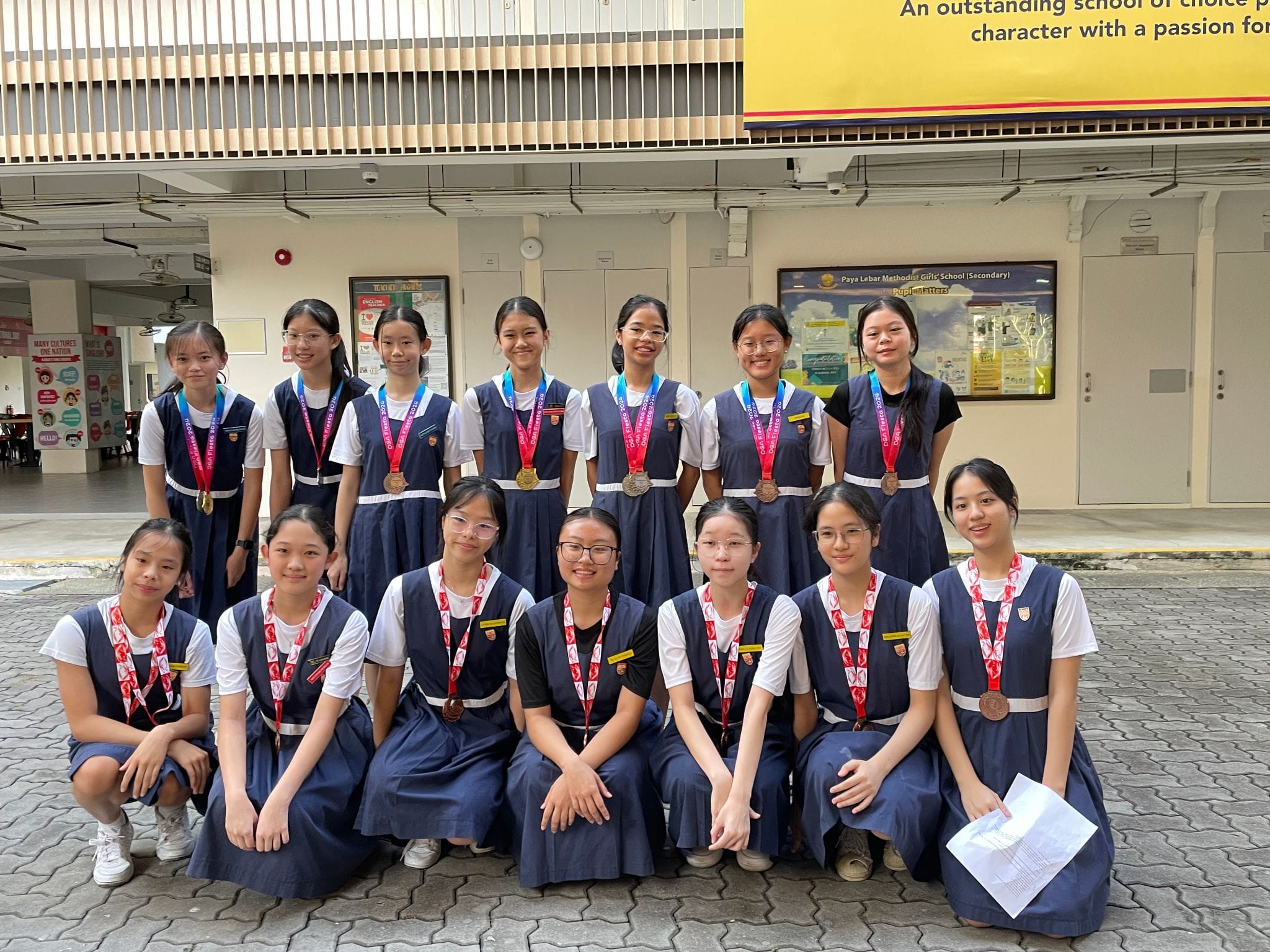 Group of schoolgirls in navy uniforms posing with medals outside Paya Lebar Methodist Girls School.