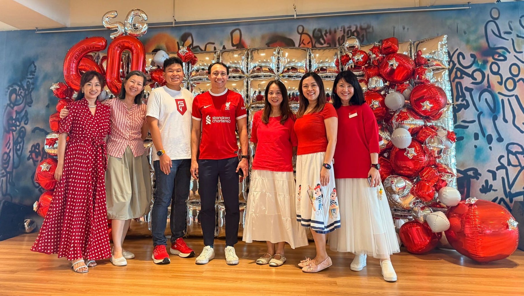 Seven people in red and white outfits pose in front of red and silver balloons spelling SG60.