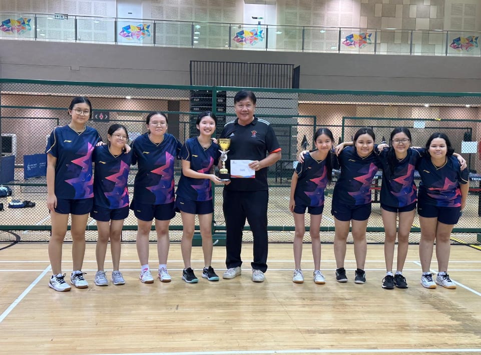 A coach and seven girls in matching sports uniforms pose together on a badminton court holding a trophy.