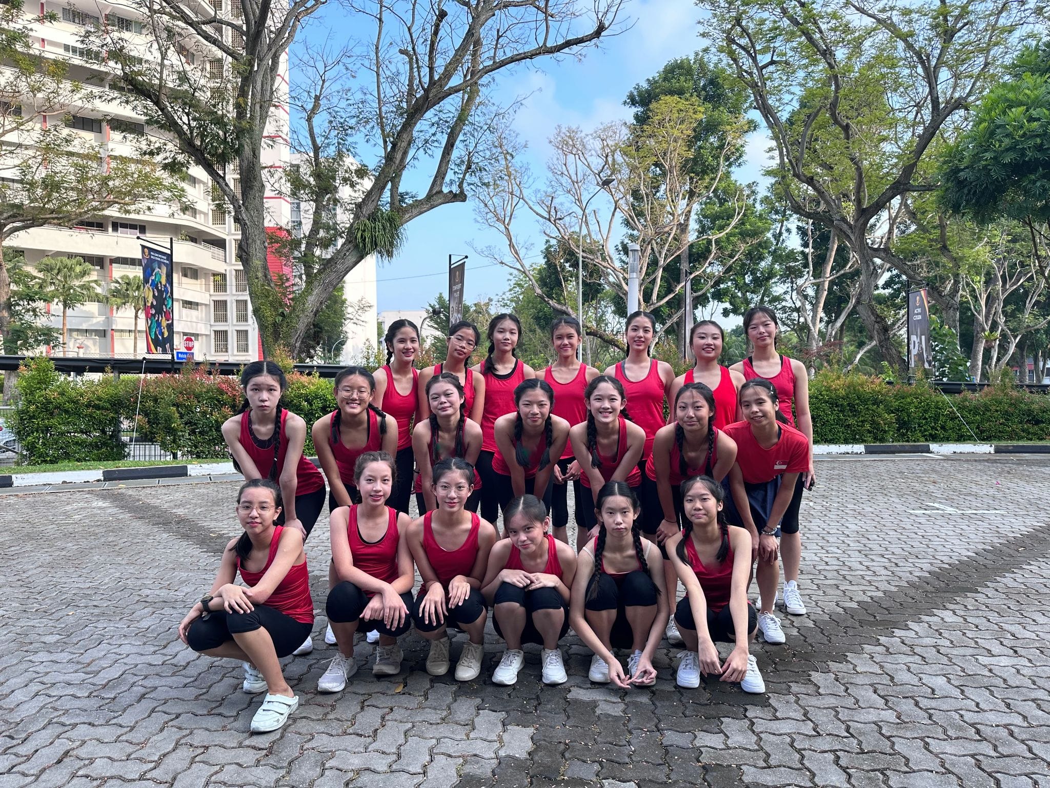 A group of young girls in red tops and black shorts pose together outdoors on a cobblestone area with trees behind them.