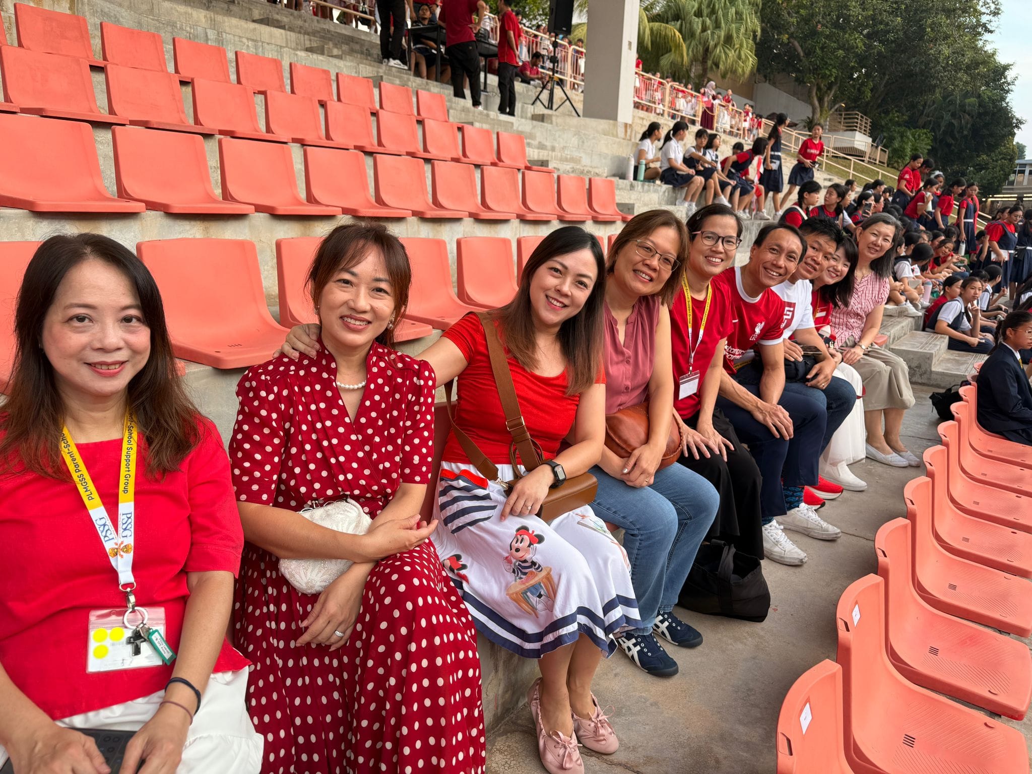 A group of smiling adults in red outfits sit together on orange stadium bleachers surrounded by a crowd.