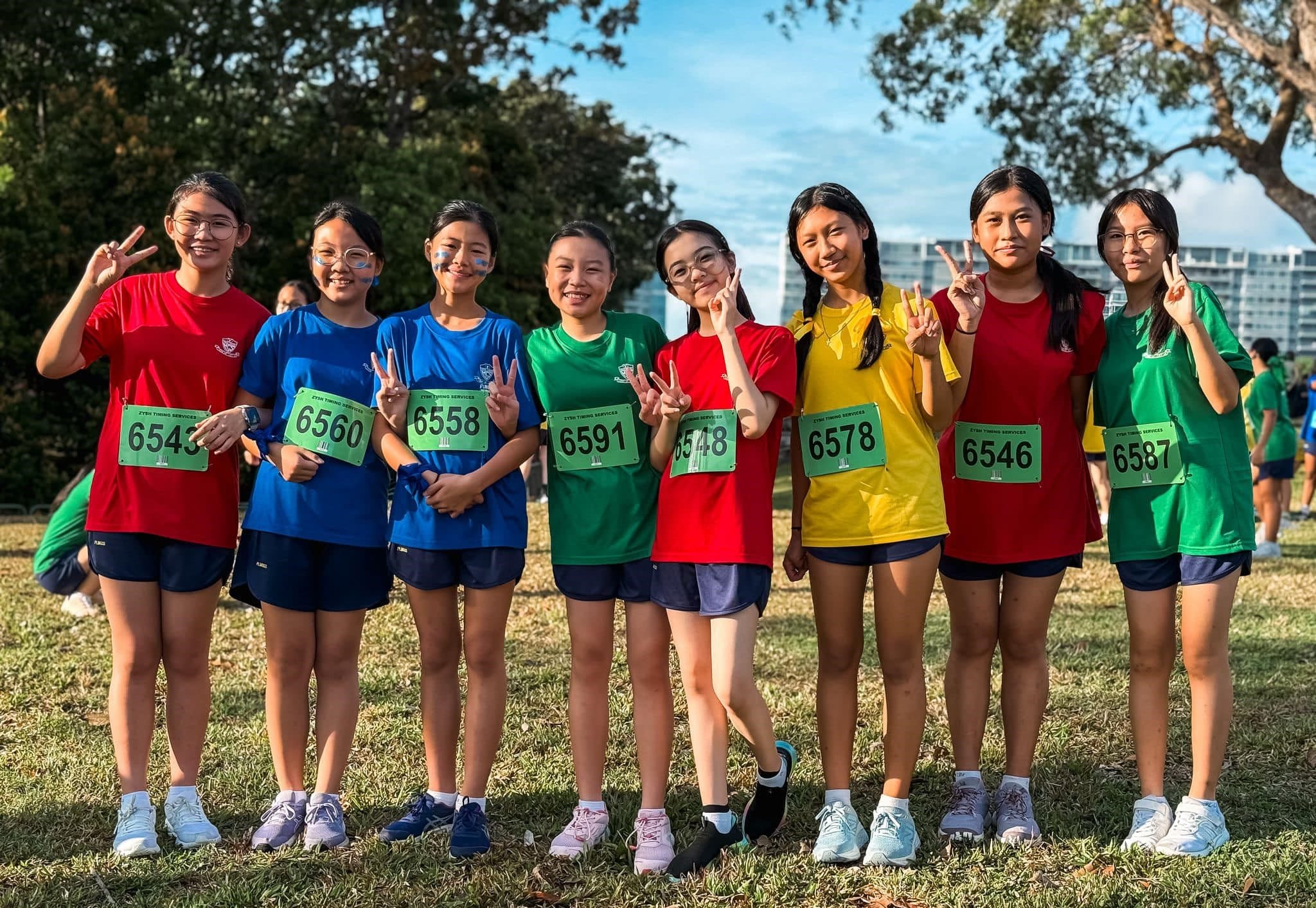 Eight young girls in colorful race shirts with green numbered bibs pose outdoors making peace signs.