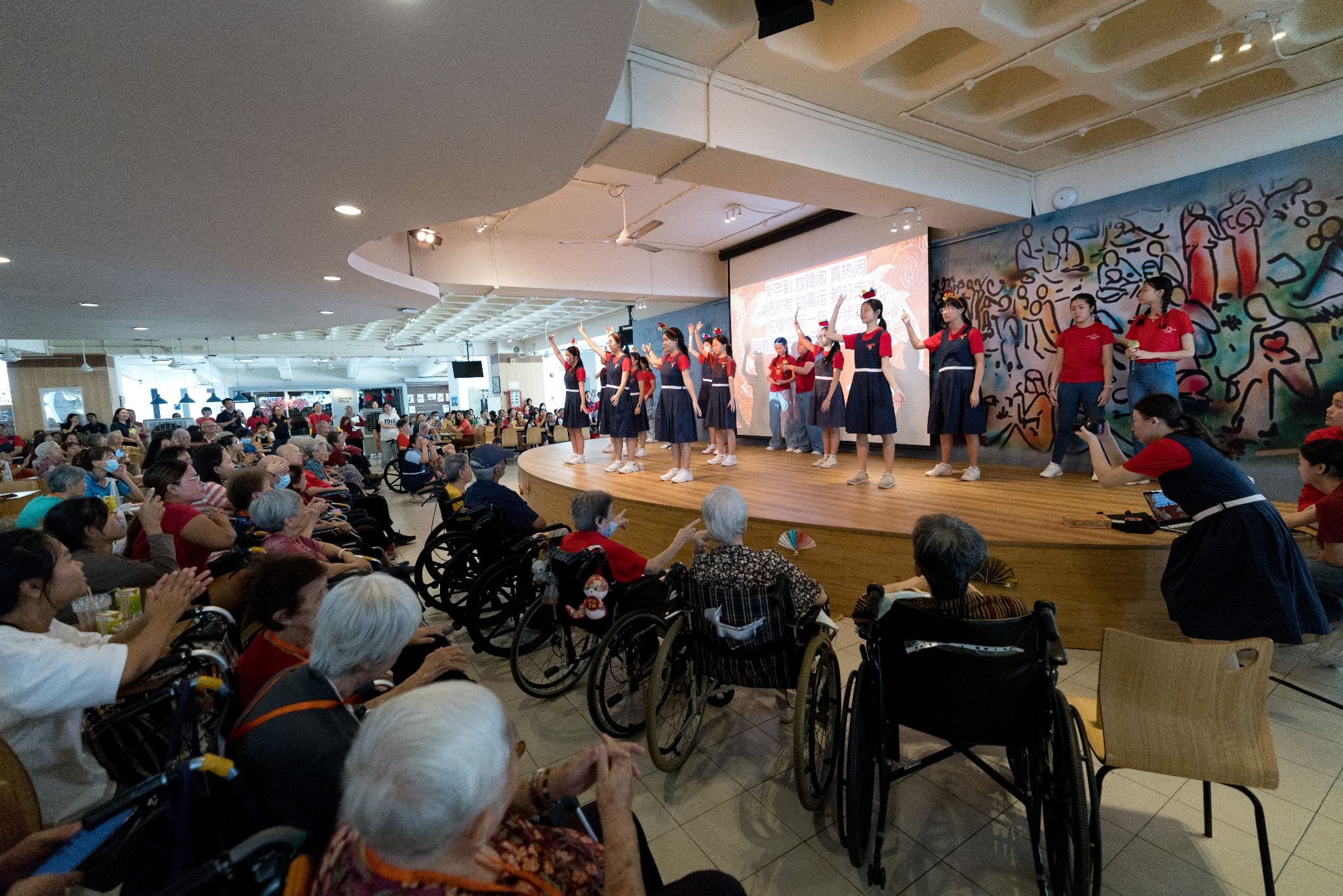 Young performers in red tops dance on a stage before a large audience of elderly people, some in wheelchairs.