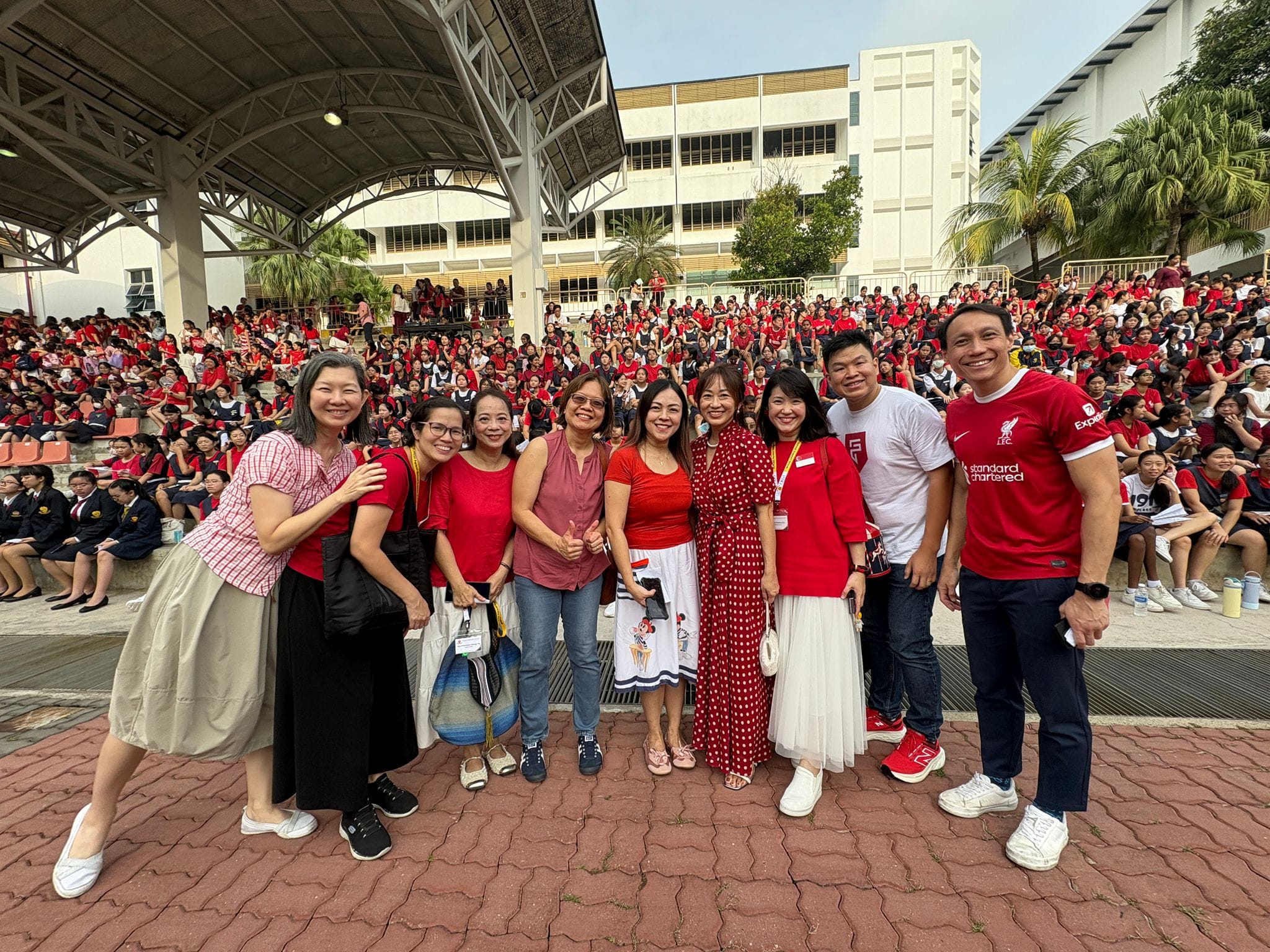 Eight adults in red outfits pose smiling outdoors, with hundreds of students in red uniforms seated on bleachers behind them.