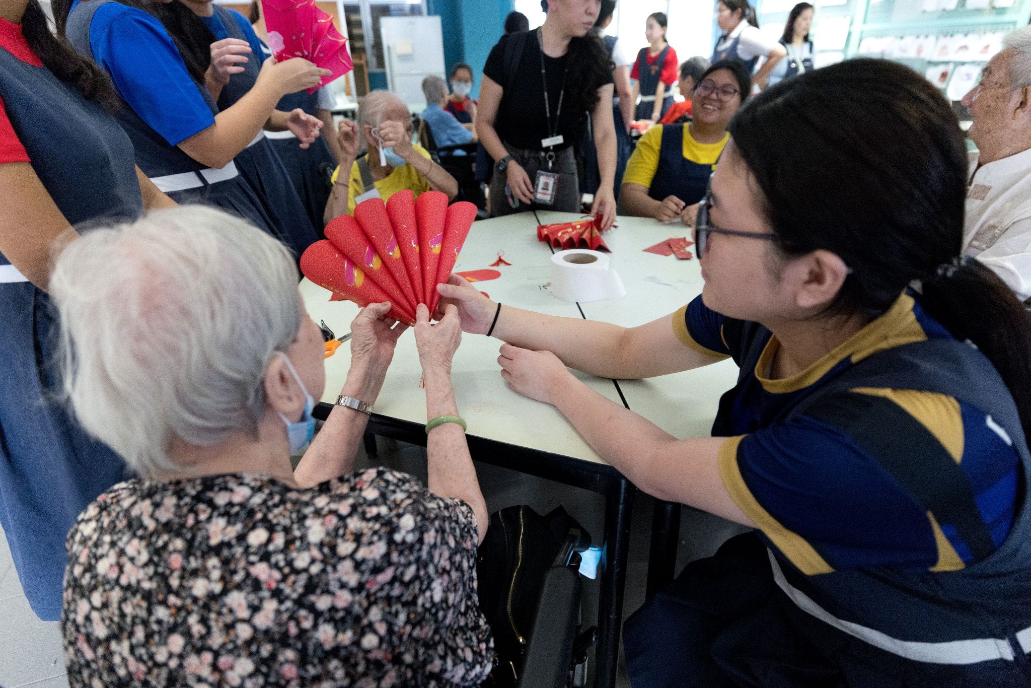 An elderly woman and a young caregiver hold a red paper fan craft together at a table with a group around them.