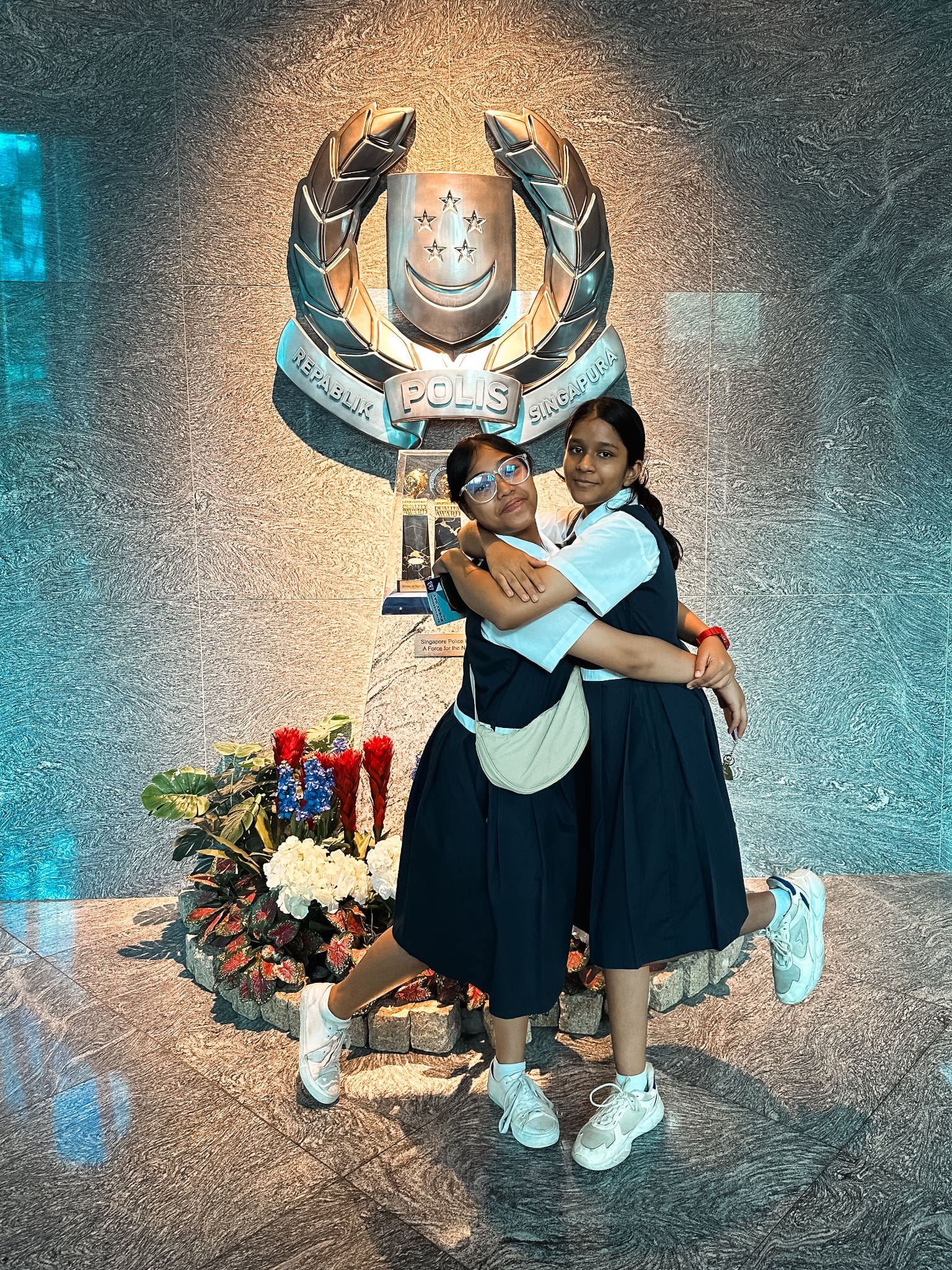 Two girls in navy uniforms hugging in front of the silver Polis Singapura crest on a textured wall.