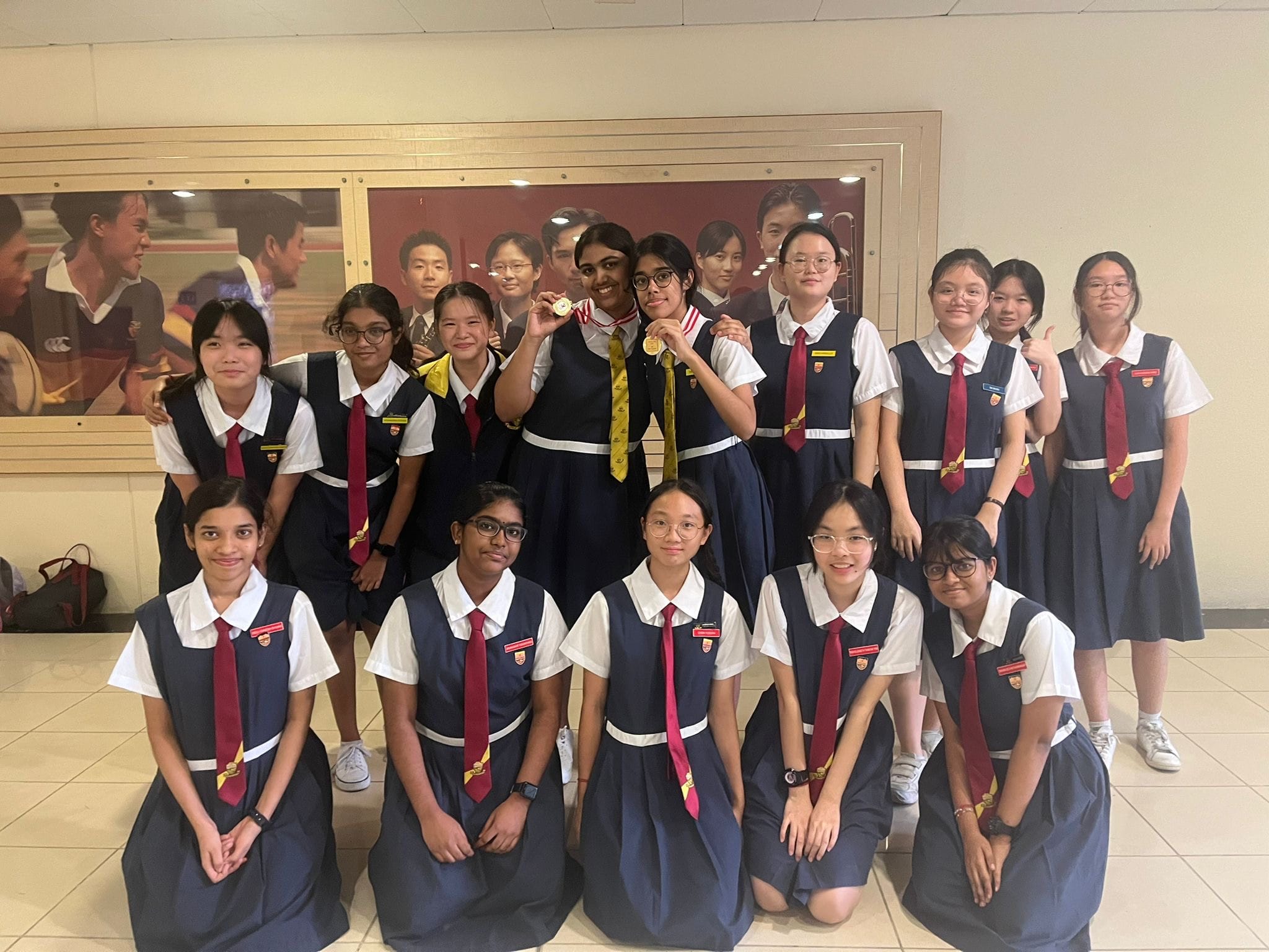 Group of teenage girls in navy school uniforms with red ties posing together, one holding gold medals in the center.