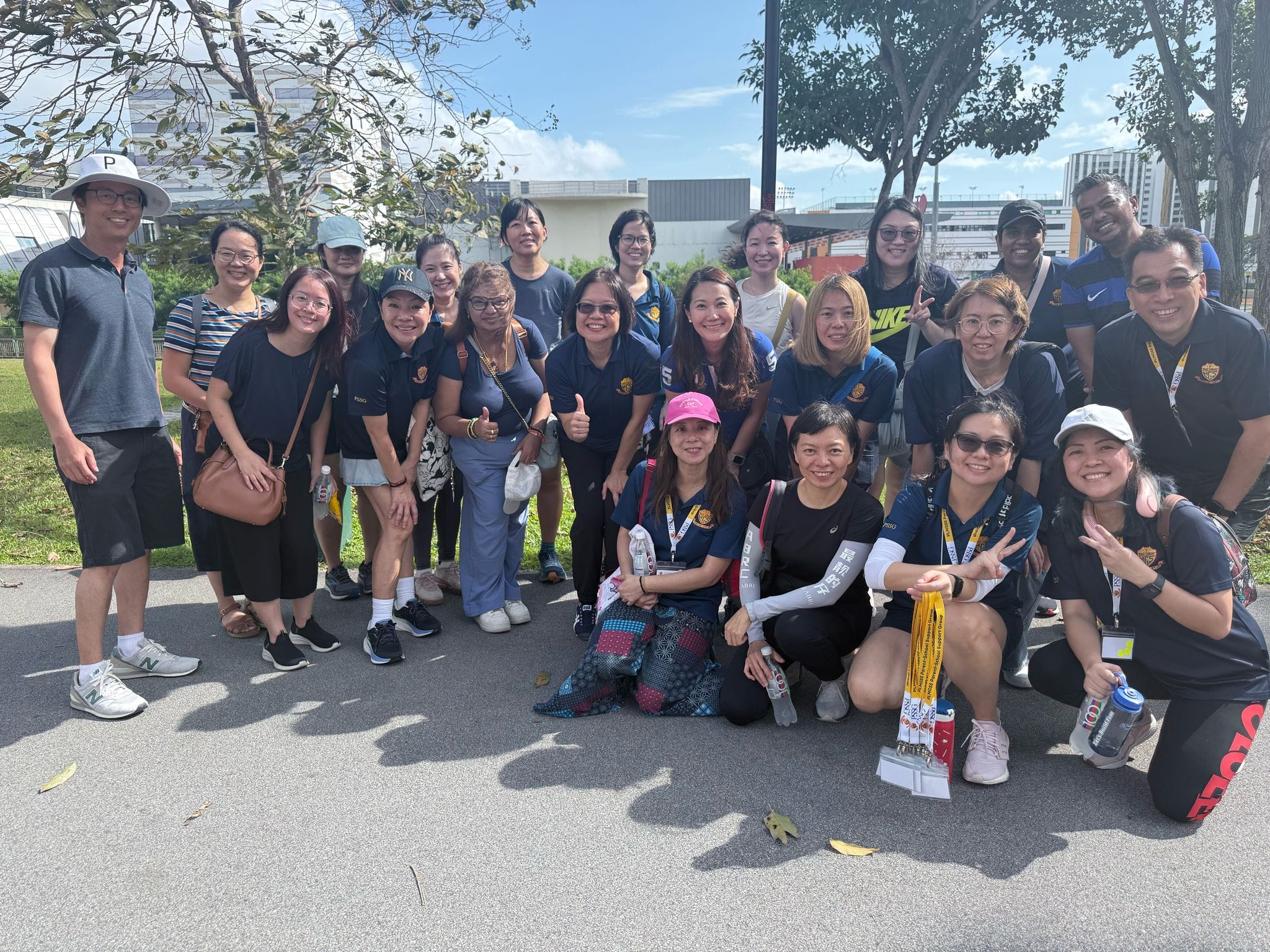 A large group of smiling adults in navy shirts pose together outdoors on a sunny day near trees and buildings.