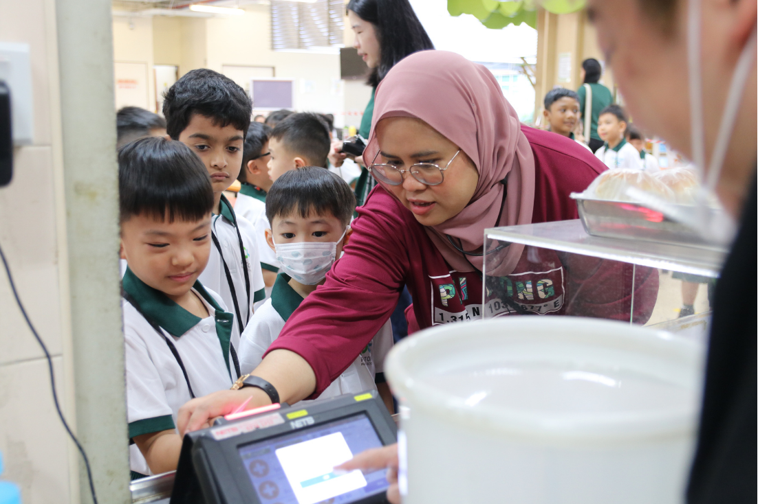 Woman wearing a pink hijab helps young students use a touchscreen device at a food counter.