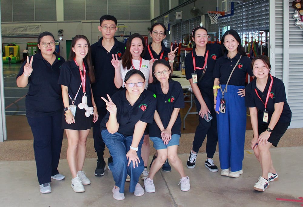 Group of ten people in black shirts posing indoors, several flashing peace signs; gym equipment behind.