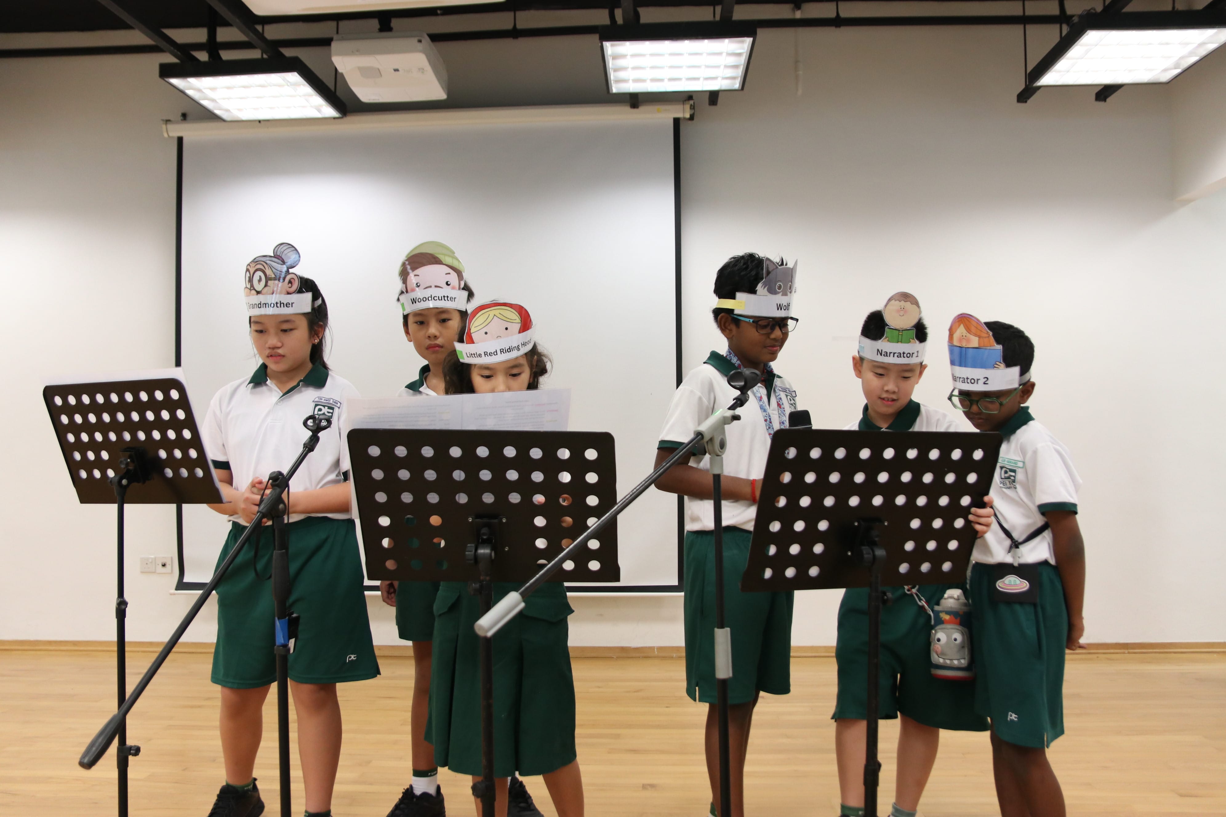 Six students participating in a storytelling session on stage, as they perform and read aloud while wearing character headbands during an interactive literacy activity.