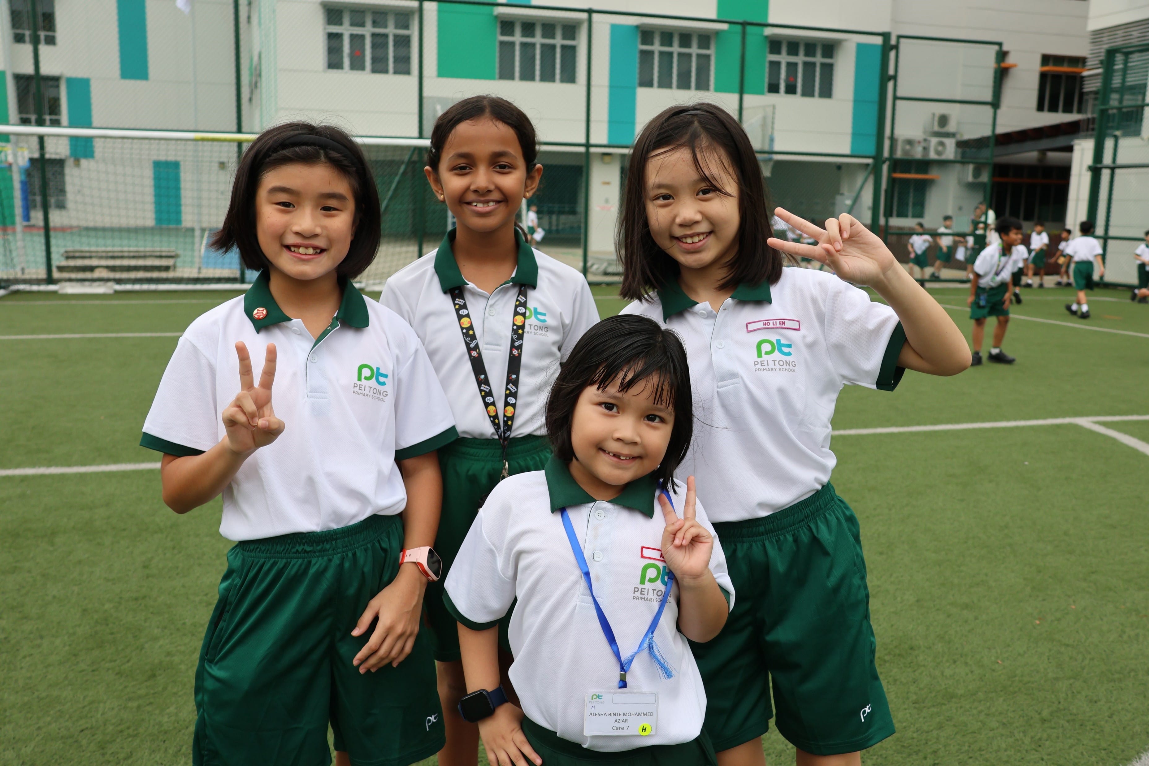 Two Primary 1 students and their Primary 4 buddies smile and pose with peace signs on the school field.