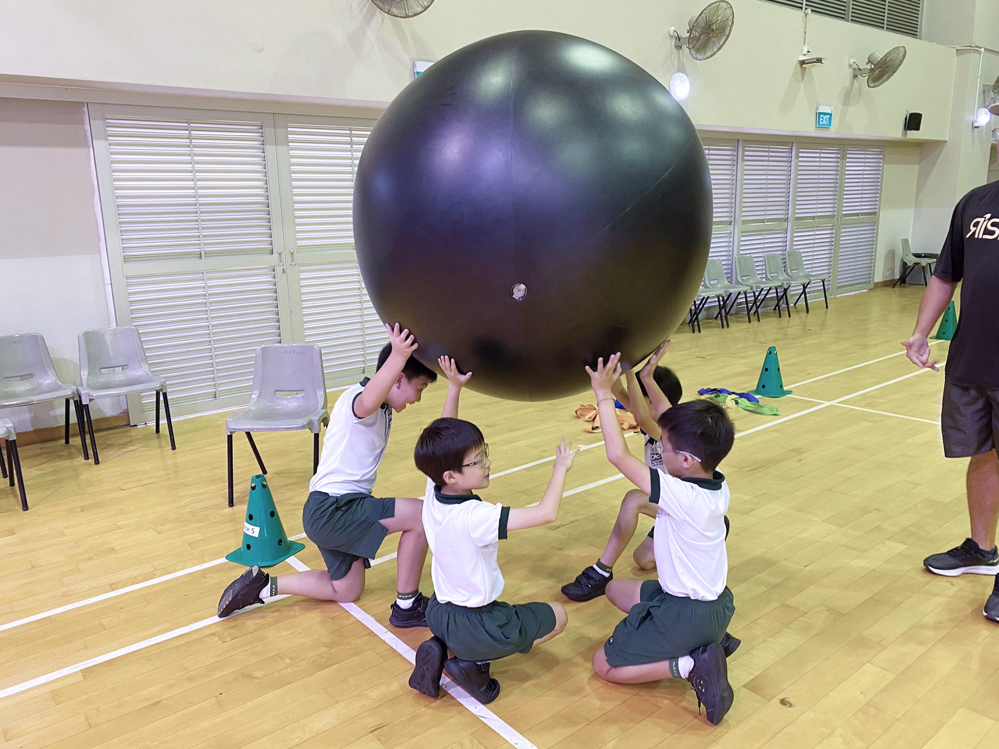 Students kneel on the sports hall floor with their hands raised as they support and balance a giant black Kin-Ball during a teamwork activity.