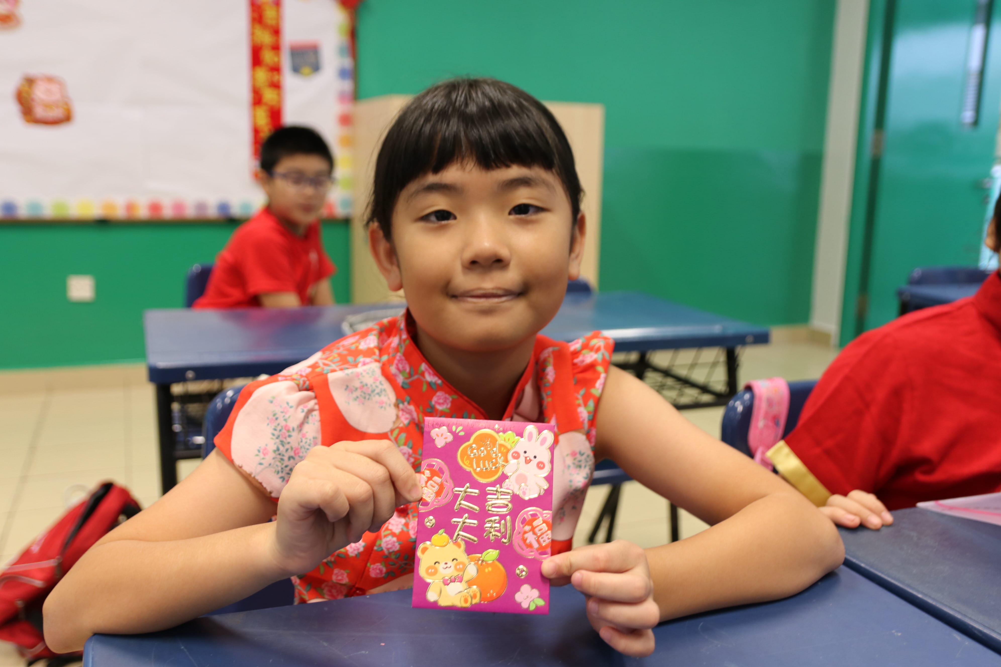A student proudly displays a Chinese New Year red packet while participating in festive classroom activities.