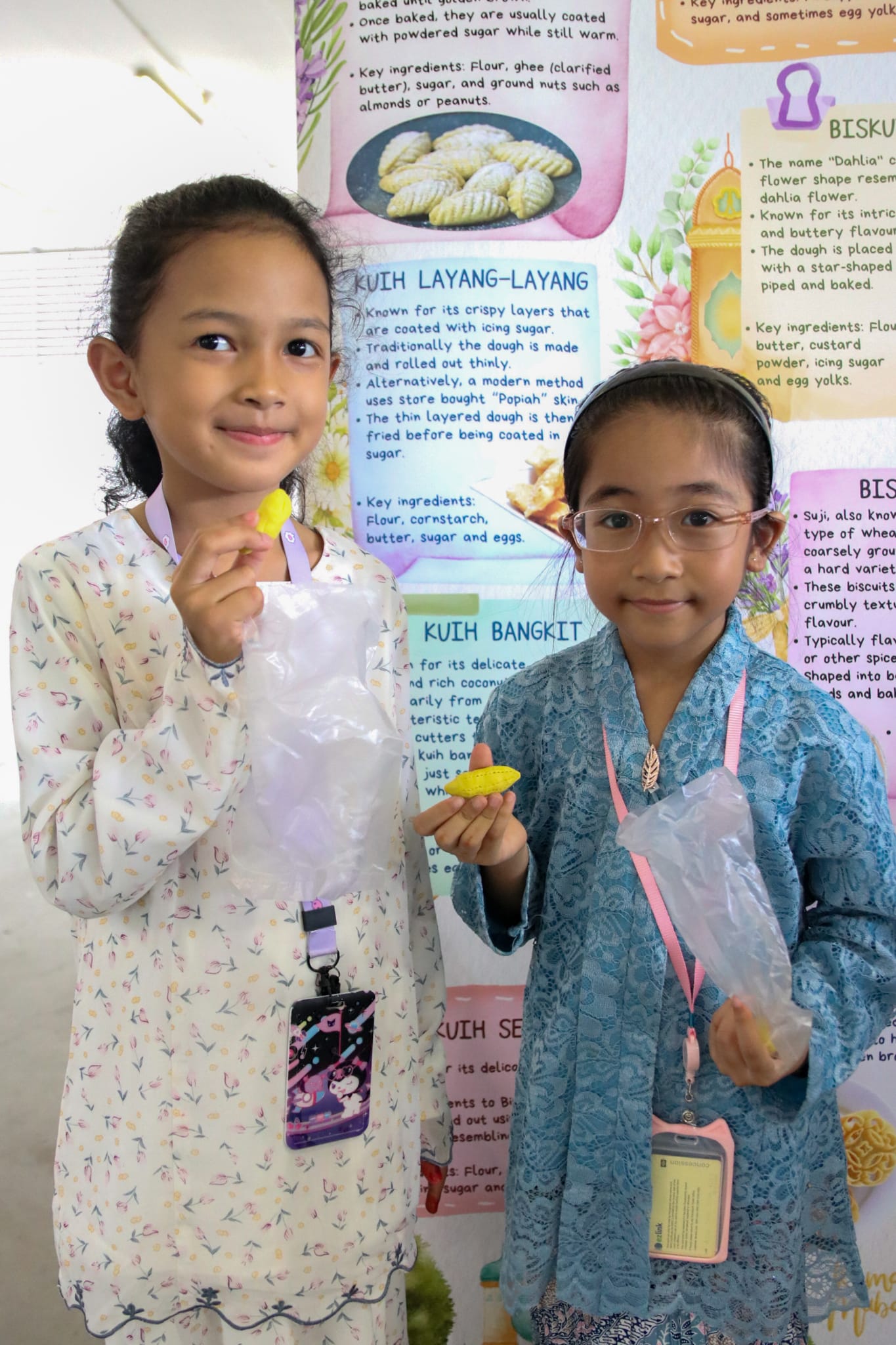 Two students in traditional attire sample Hari Raya festive cookies while exploring a display about traditional Malay delicacies.