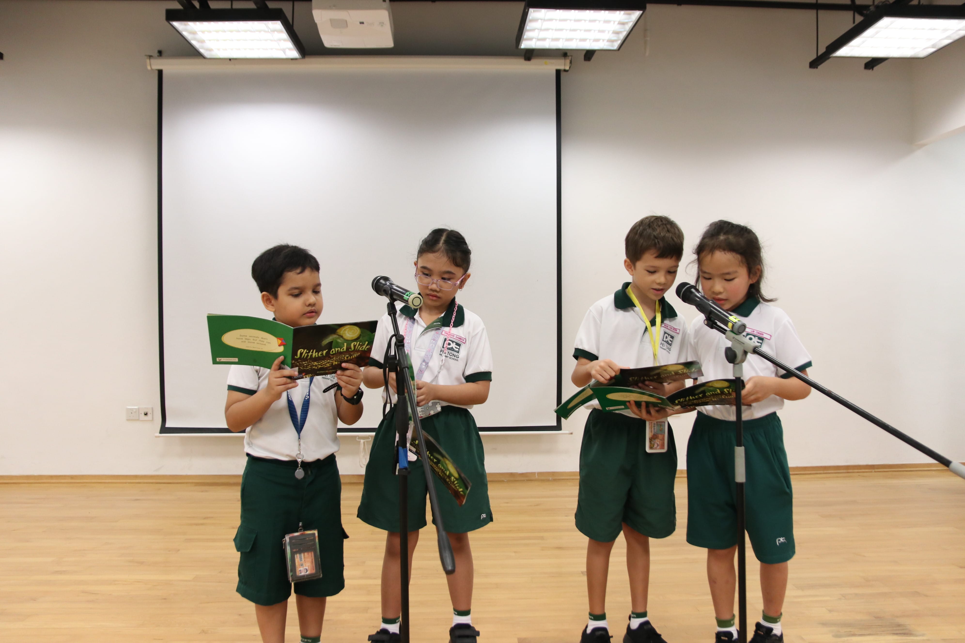 Four lower primary students reading aloud during a storytelling session, as they take turns presenting and performing a story using microphones on stage.