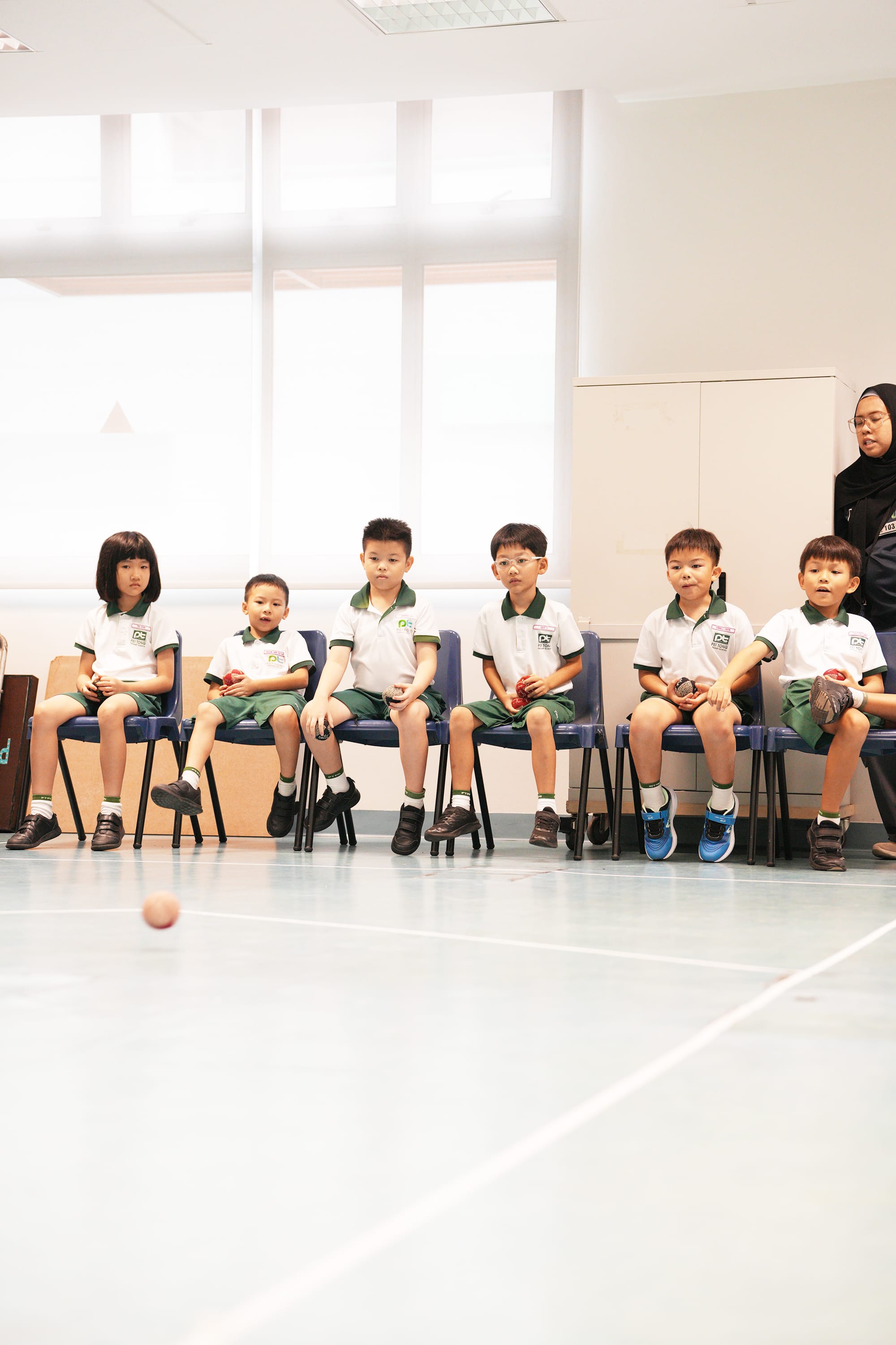 Young students sit in a row, watching attentively and waiting for their turn to participate in an indoor ball activity during the Sports Carnival.