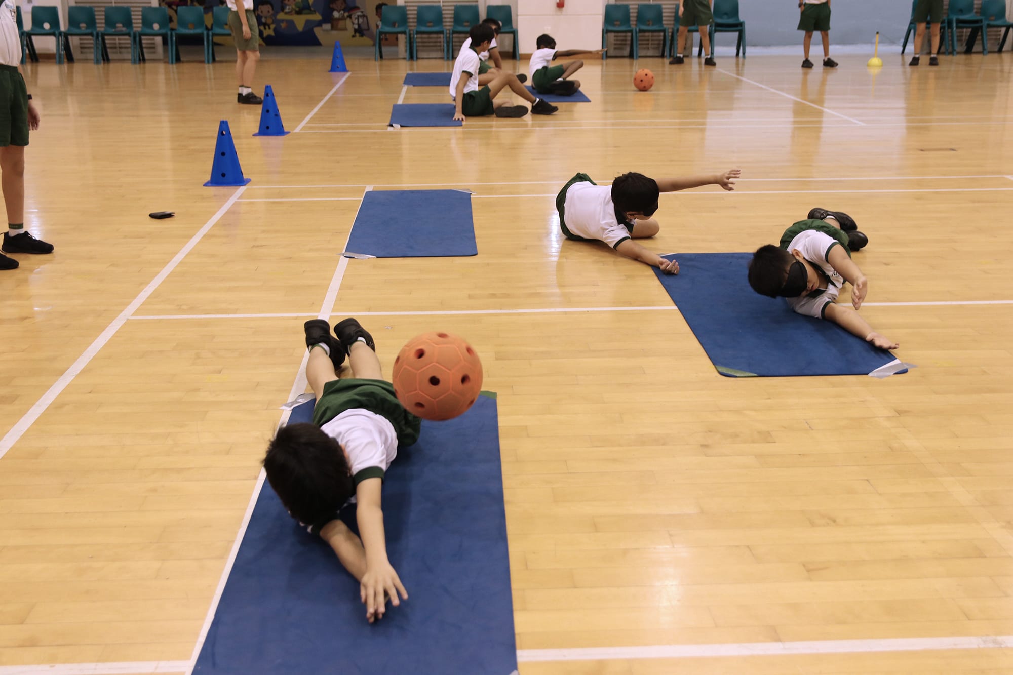 Students lie on blue exercise mats in an indoor sports hall while reaching out to stop or catch an orange ball rolling across the floor during a GOA activity. Cones and additional mats are arranged around the playing area, with other students waiting for their turn in the background.