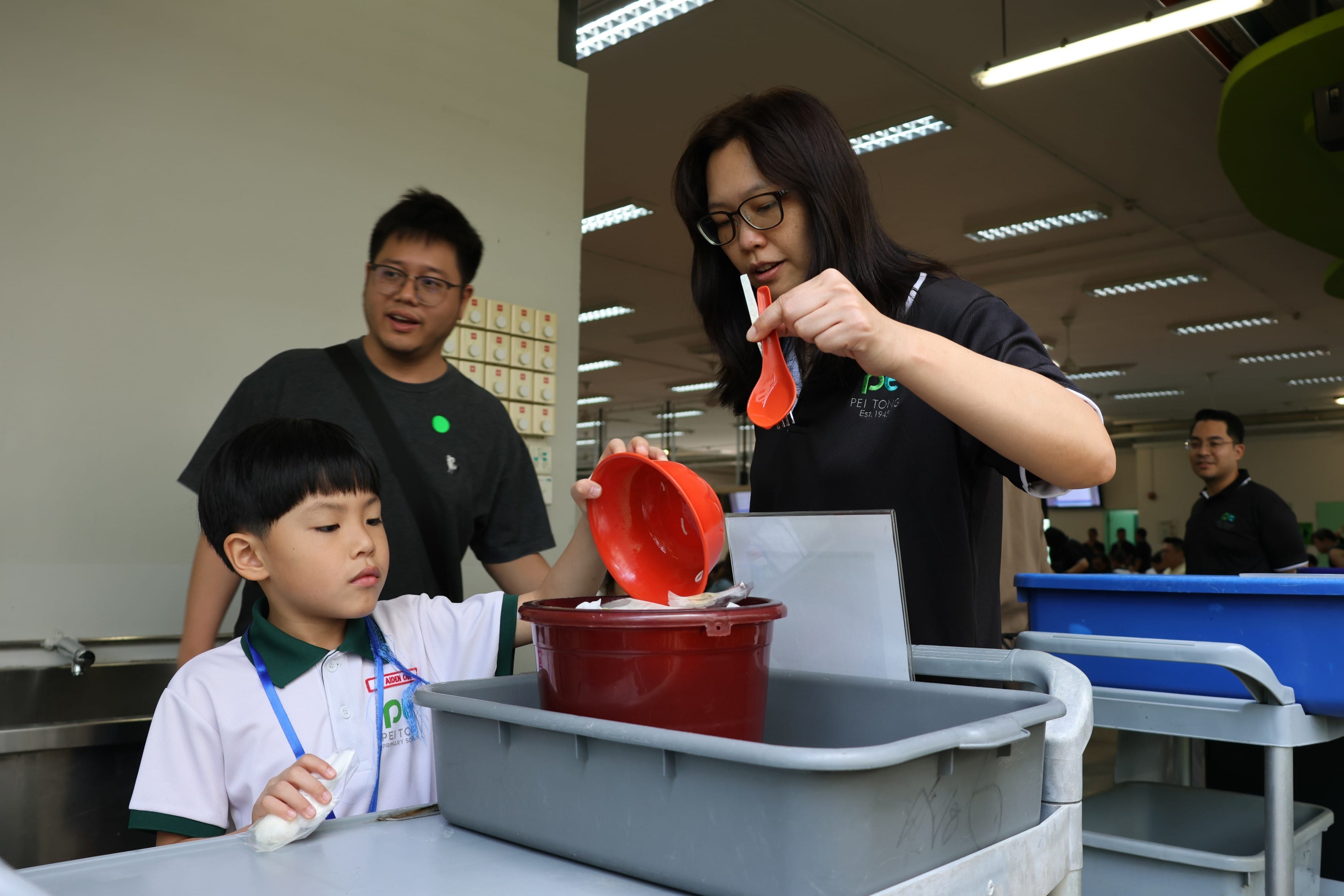 Teacher guides a Primary 1 boy as he pours food scraps into a red bucket on a tray-return cart.