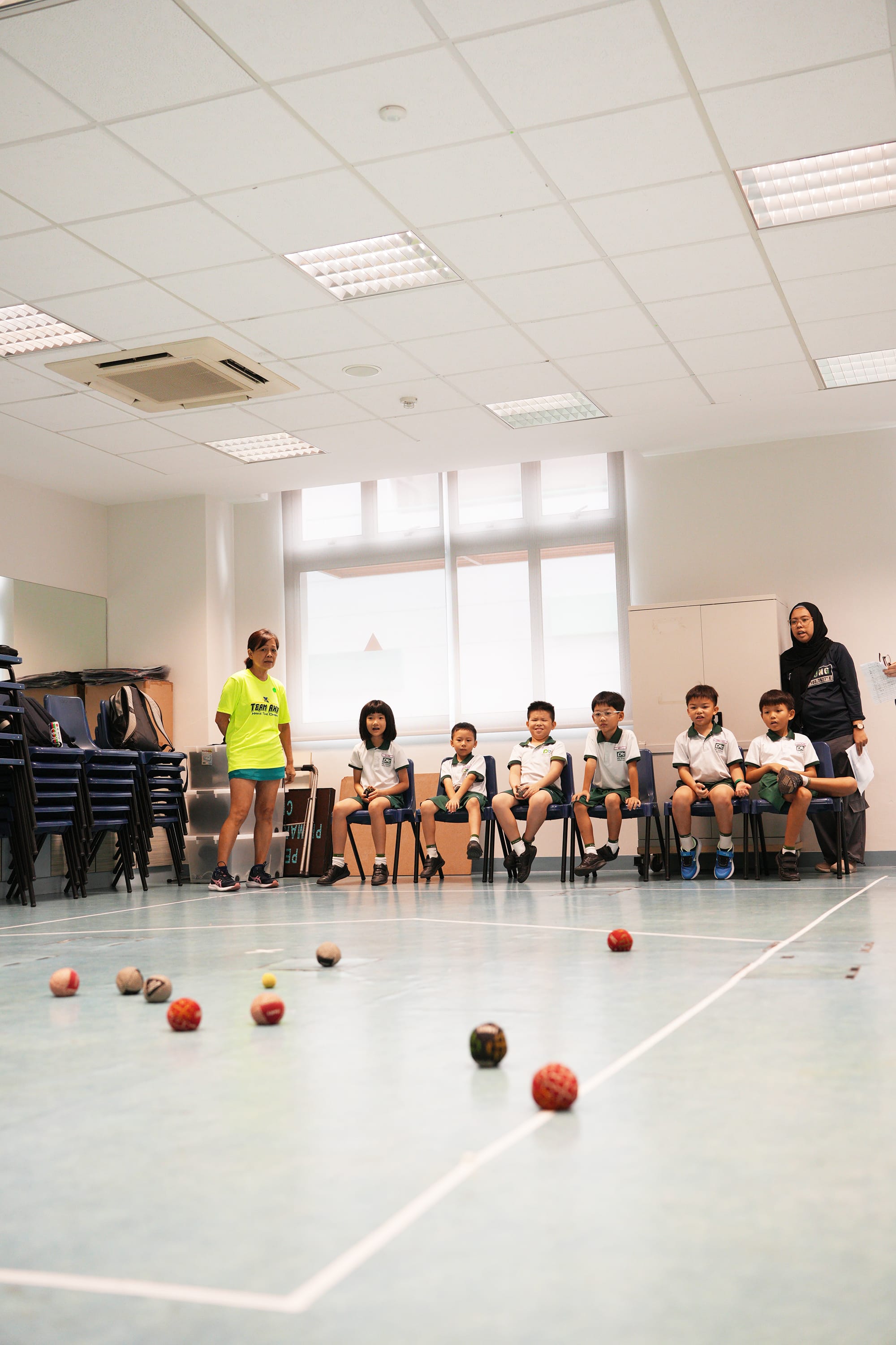Students sit in a row of chairs watching several coloured Boccia balls spread across the indoor court as an instructor stands beside them, guiding the activity before the next turn.