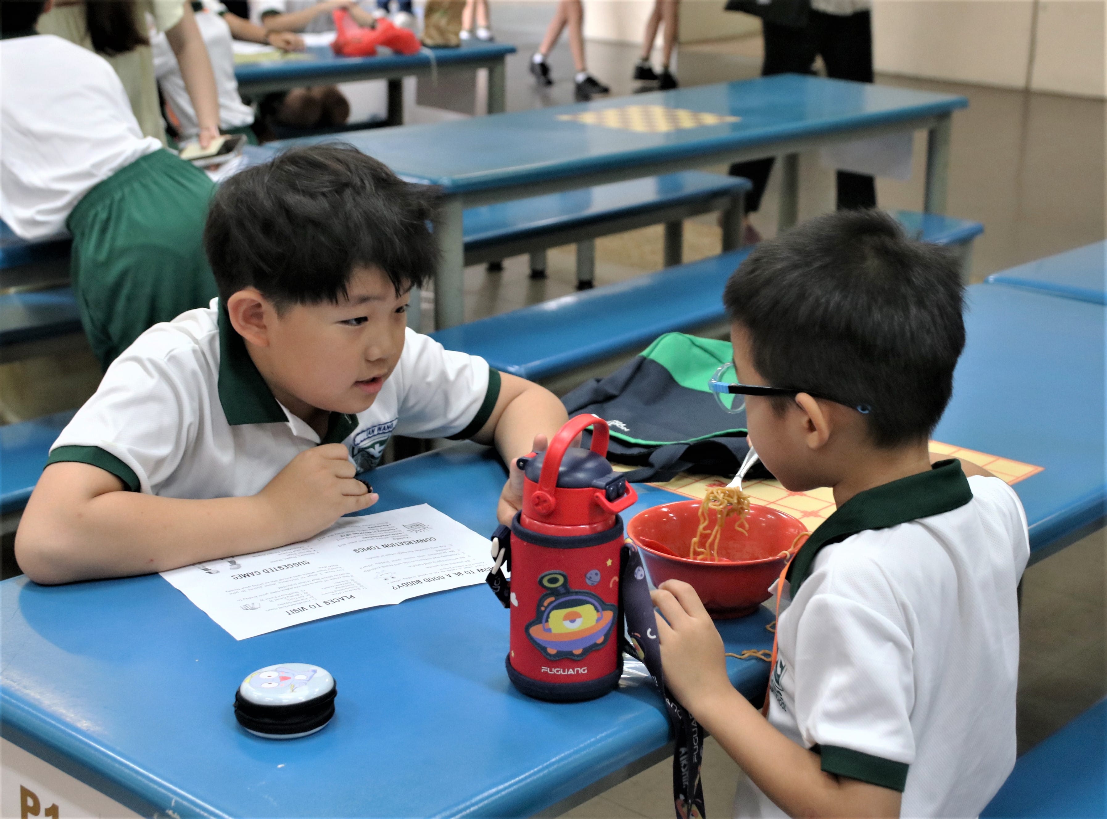 Primary 4 buddy and Primary 1 student sit across from each other at a blue canteen table, talking while the younger student eats a bowl of noodles.