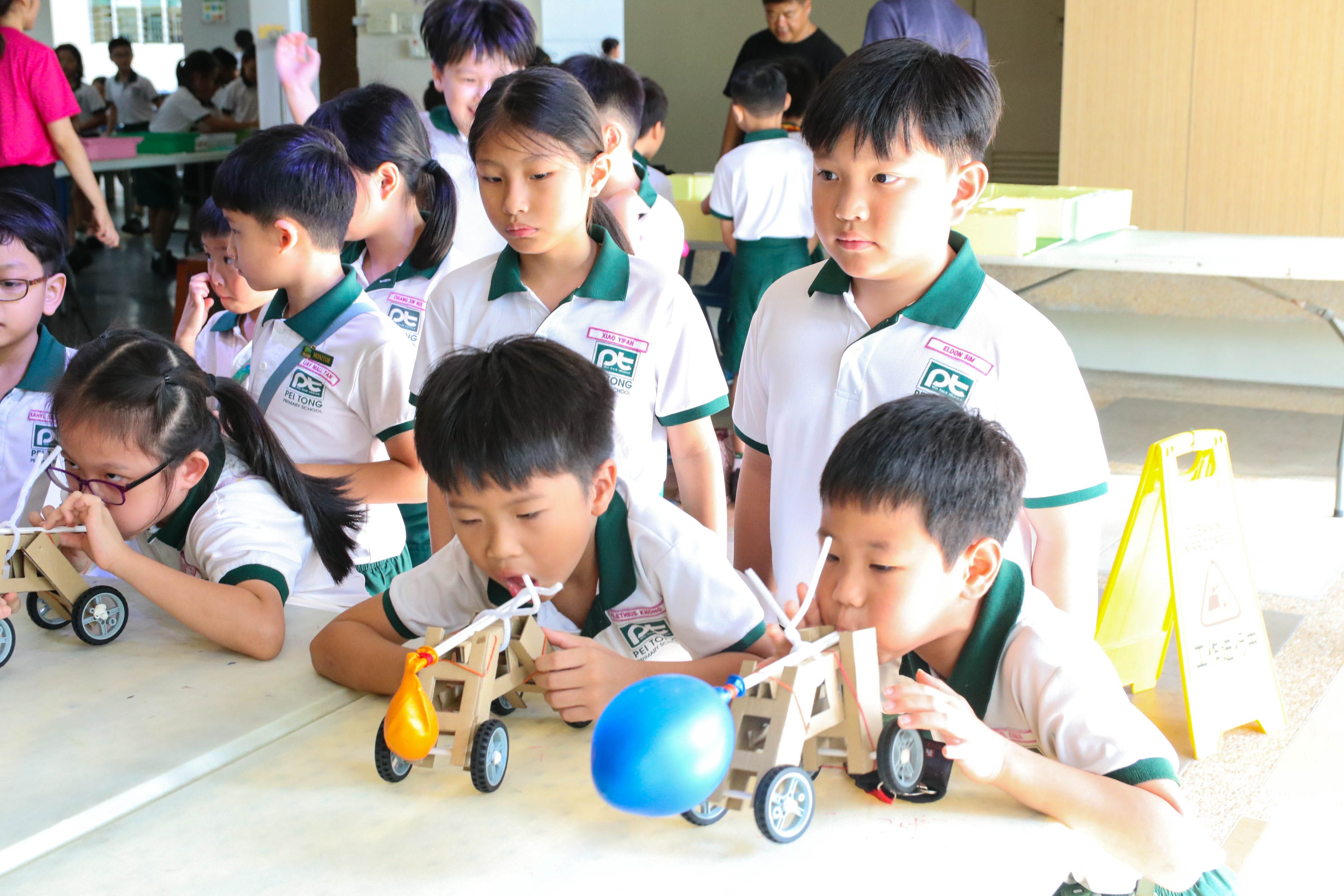 Students testing balloon-powered cars during a hands-on STEM activity, with students blowing into straws attached to balloon cars while classmates watch closely around the table.