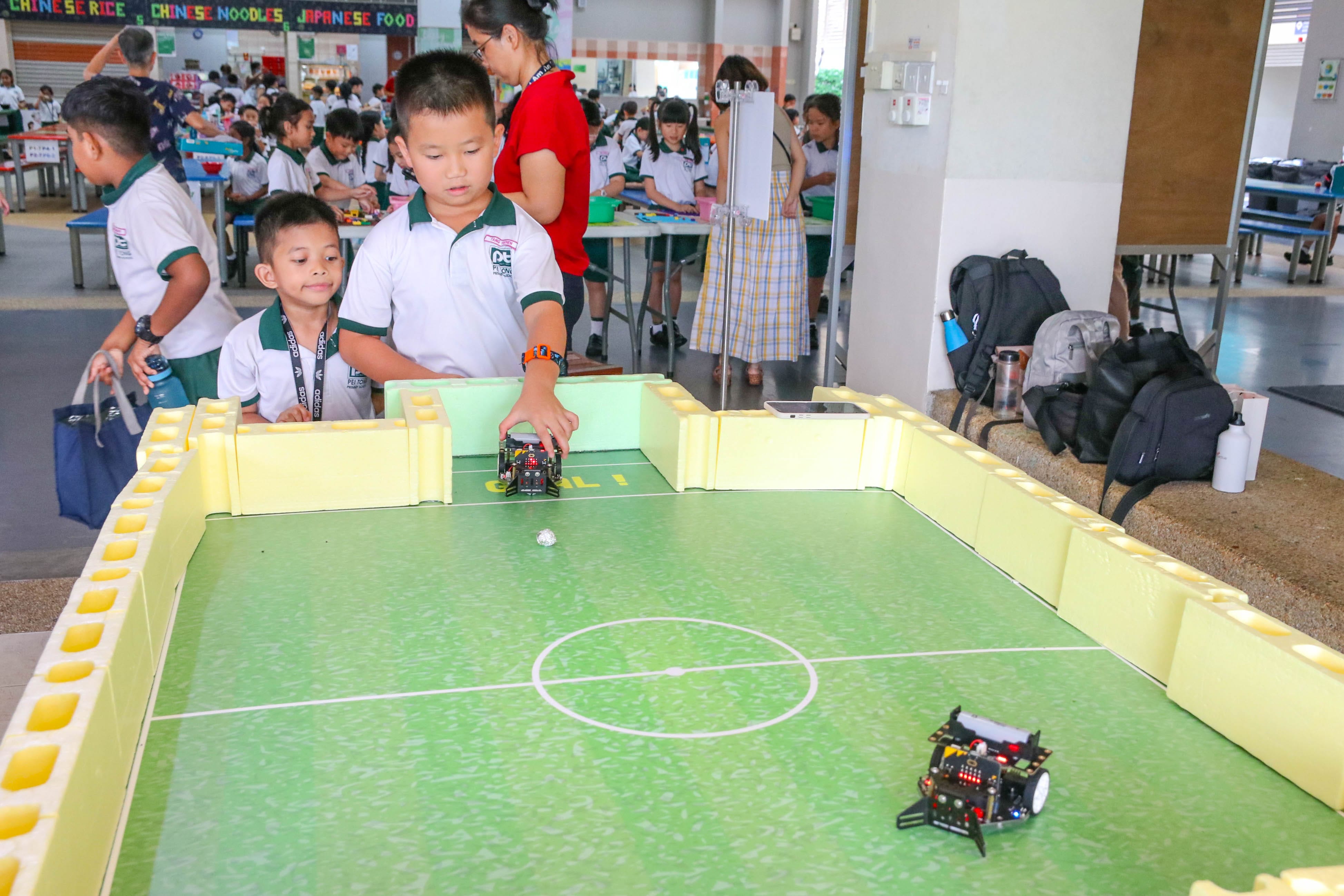 Students participating in a robotics activity, with a student controlling a small robot on a miniature football field while classmates and teachers observe in the school canteen area.
