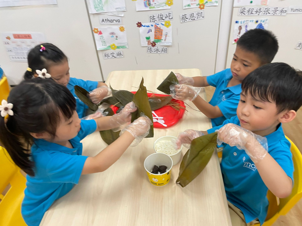 Children collaborate to wrap rice dumplings with bamboo leaves, exploring cultural traditions through hands-on learning and teamwork.
